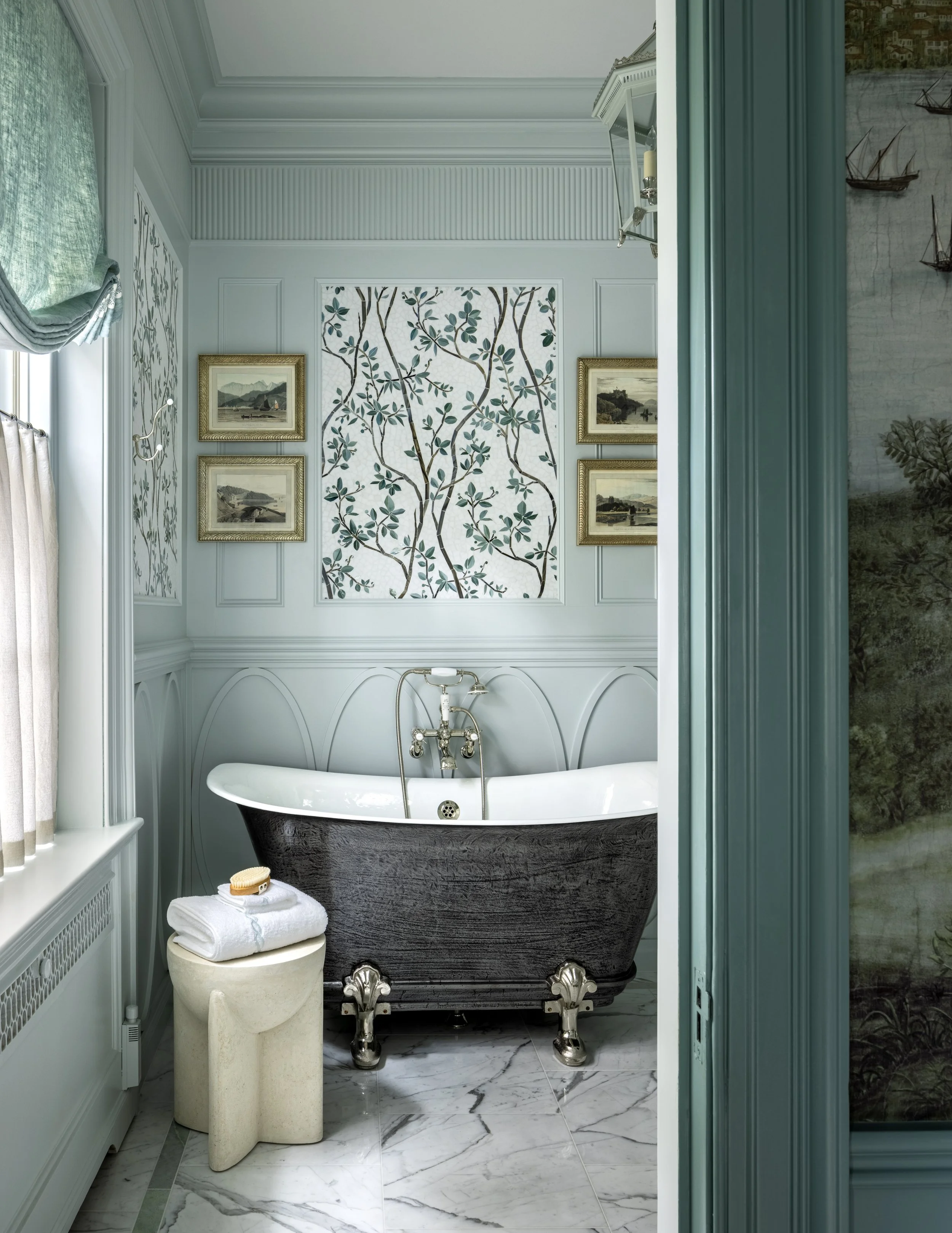 A luxurious bathroom features a vintage black clawfoot bathtub with ornate silver feet, against a wall with decorative white paneling and framed landscape paintings. A small side table holds a rolled white towel and a bath brush, and a window with li