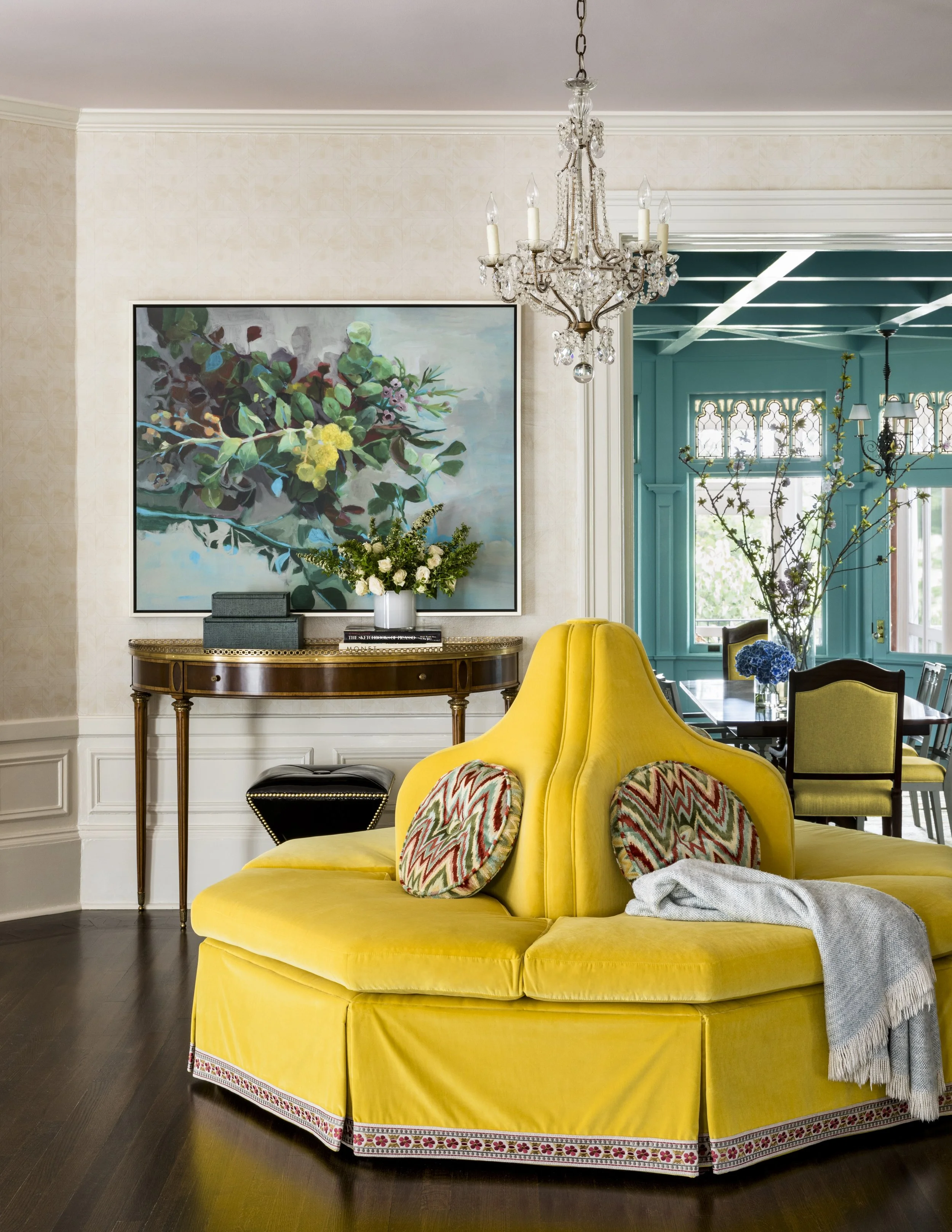 A living room corner with a vibrant yellow upholstered sofa, colorful patterned pillows, a blanket, a floral arrangement with white roses, a dark-colored table, a framed floral painting, a chandelier, and large windows with blue trim in the backgroun