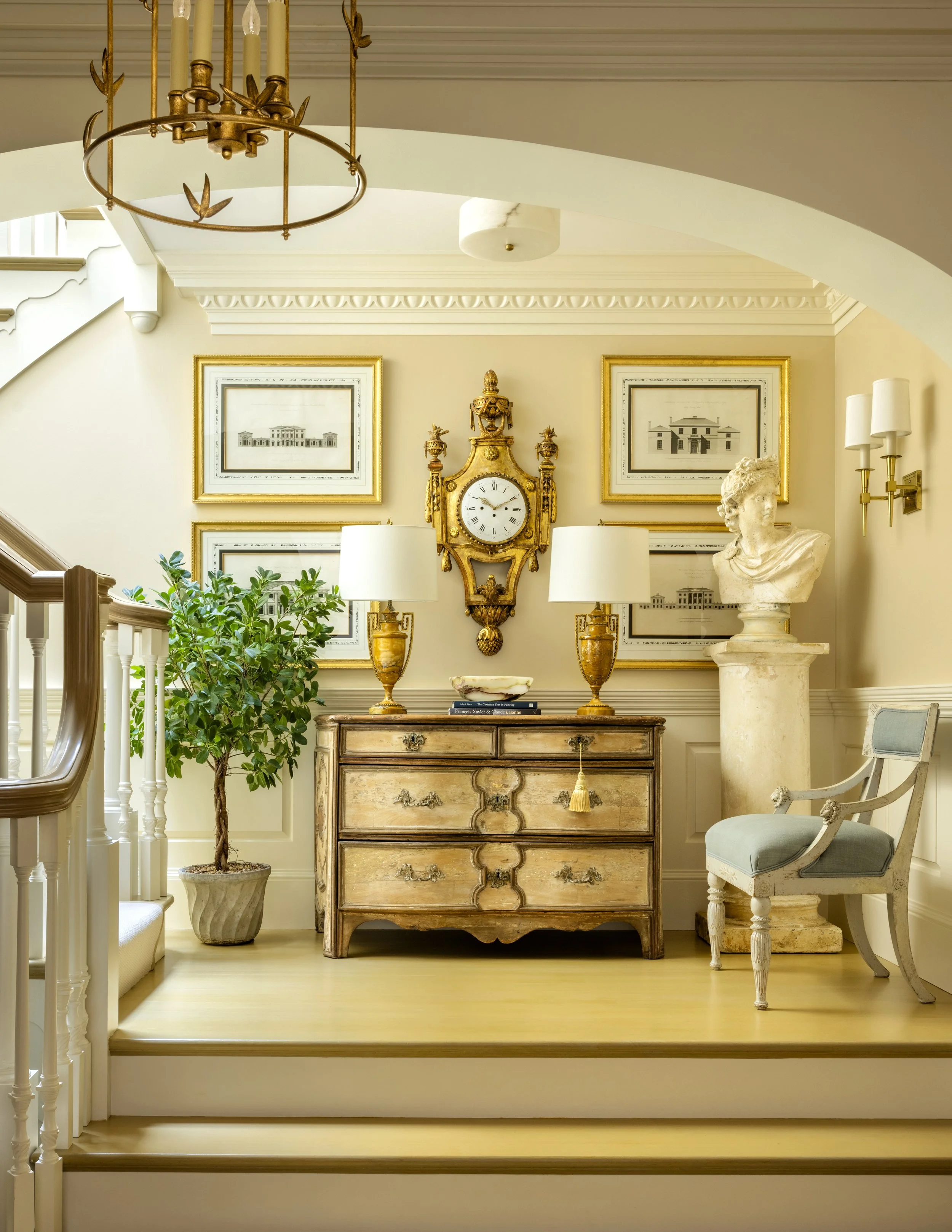 Elegant interior room with a carved wooden cabinet, a classical bust sculpture, framed architectural drawings, and a vintage gold wall clock with surrounding artwork and decorative lamps.