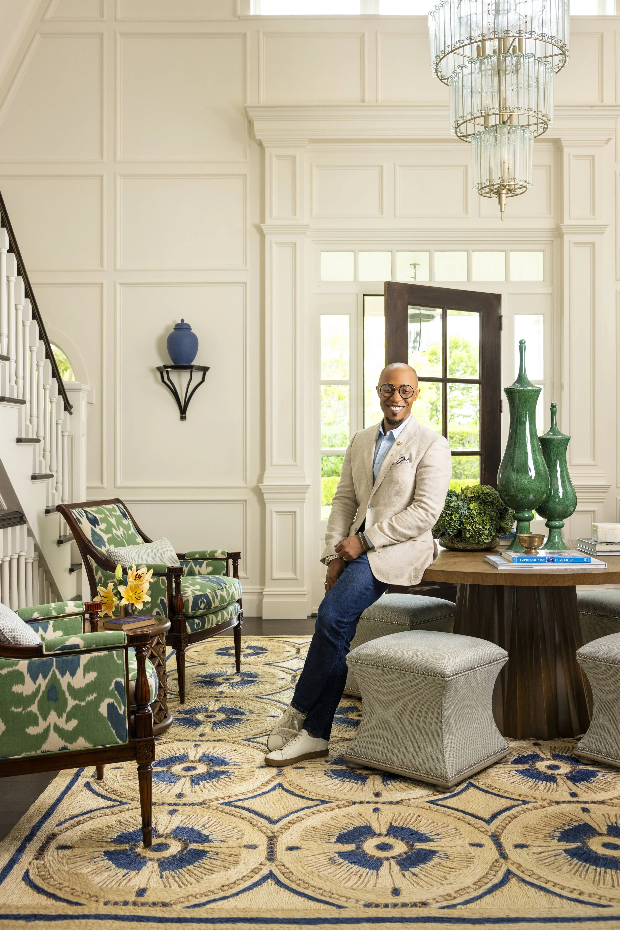 A man smiling, wearing glasses, a light-colored blazer, and jeans, standing in a well-decorated room with a large window, green decorative vases, and patterned armchairs. The room has white paneled walls, a chandelier, and a colorful rug.