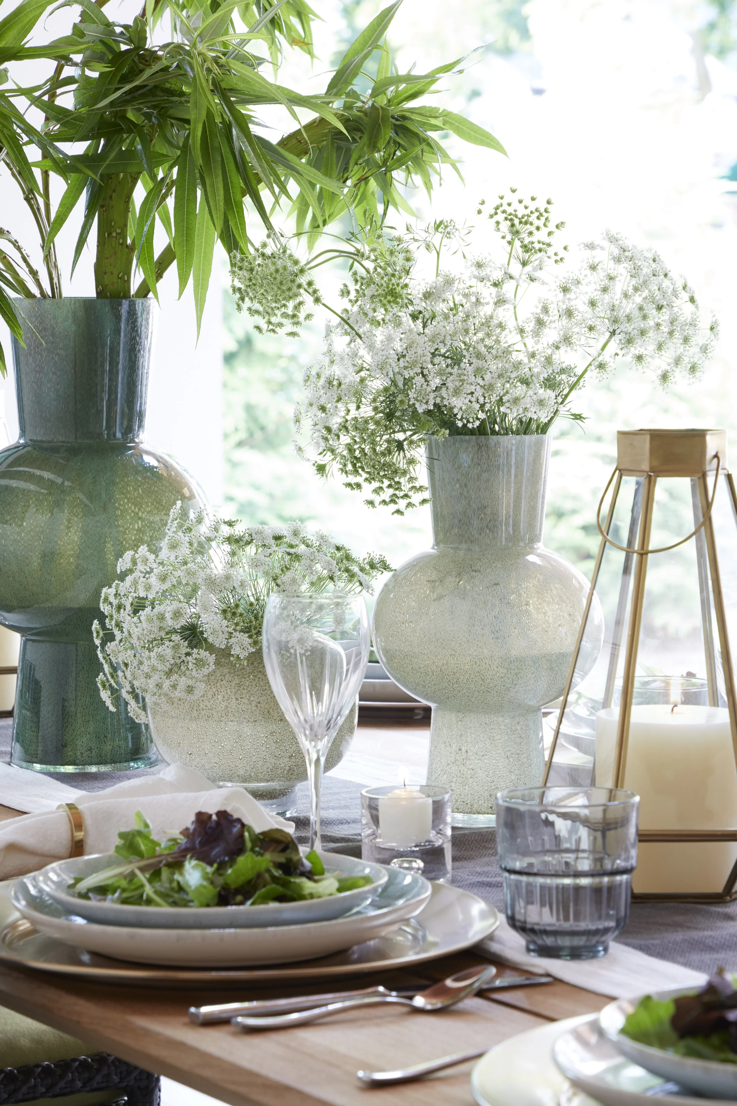A dining table setup with a salad, glassware, candles, and decorative vases filled with white flowers and green foliage, illuminated by natural light.