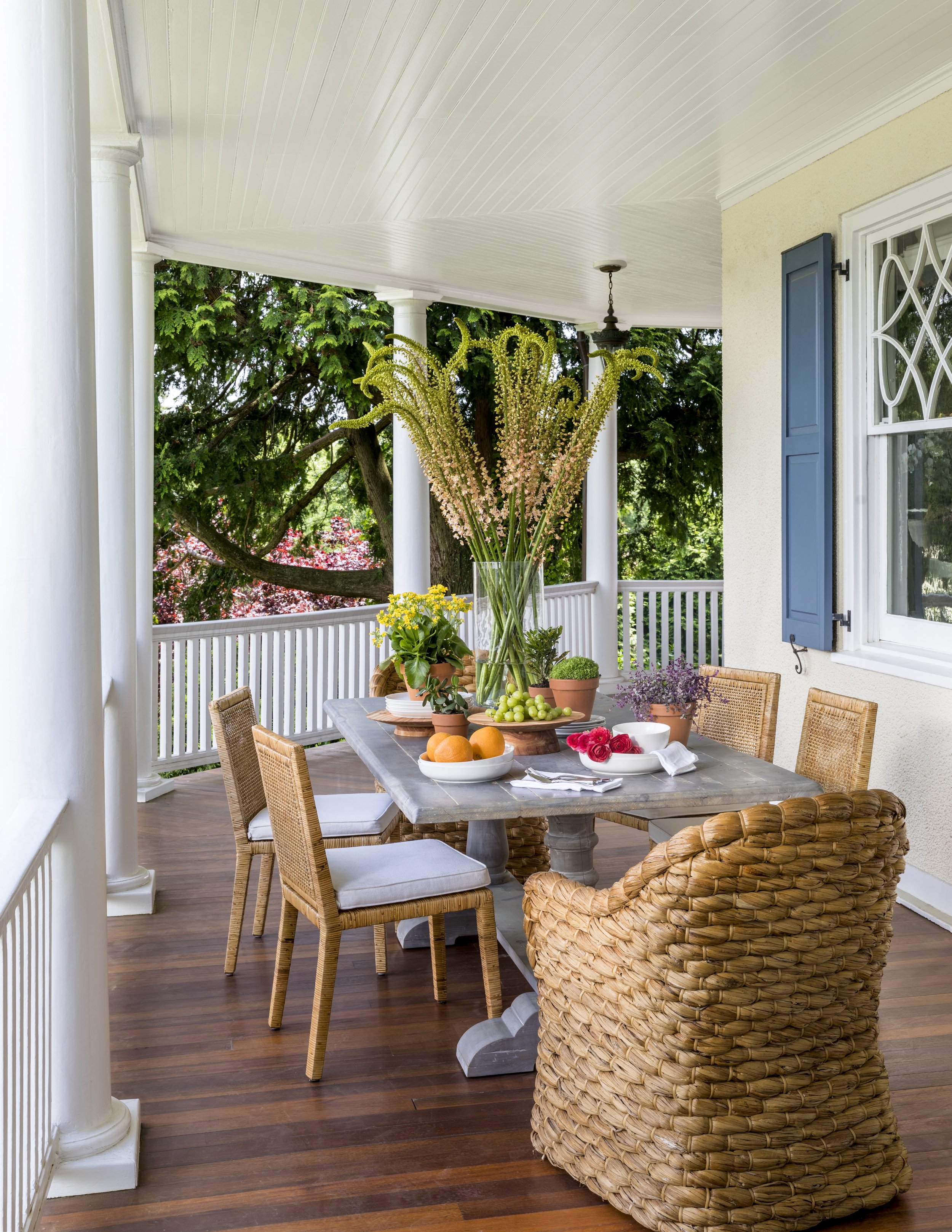 A porch with a dining table set for a meal, decorated with a large glass vase of pink and green flowers and smaller potted plants, surrounded by wicker chairs and a white railing with greenery in the background.
