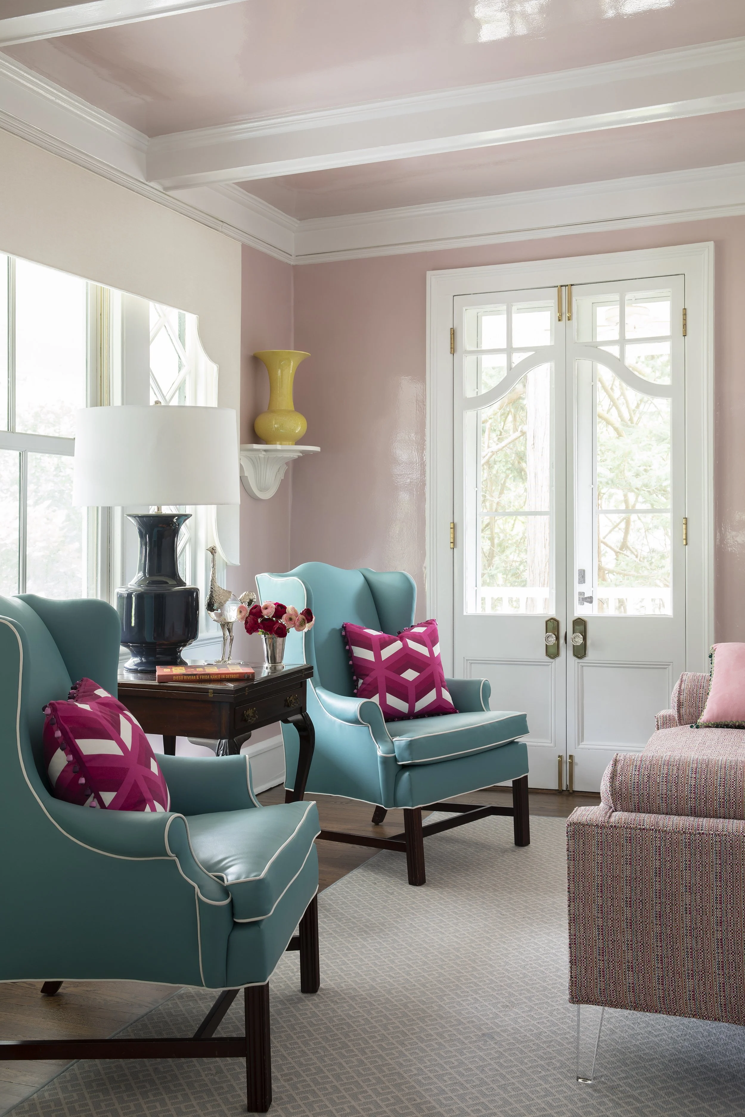 Living room with pink and beige walls, white trim, large window, turquoise armchairs with pink and white patterned pillows, side table with a black vase lamp, books, and glass vase with flowers, yellow decorative vase on wall shelf, beige carpet.