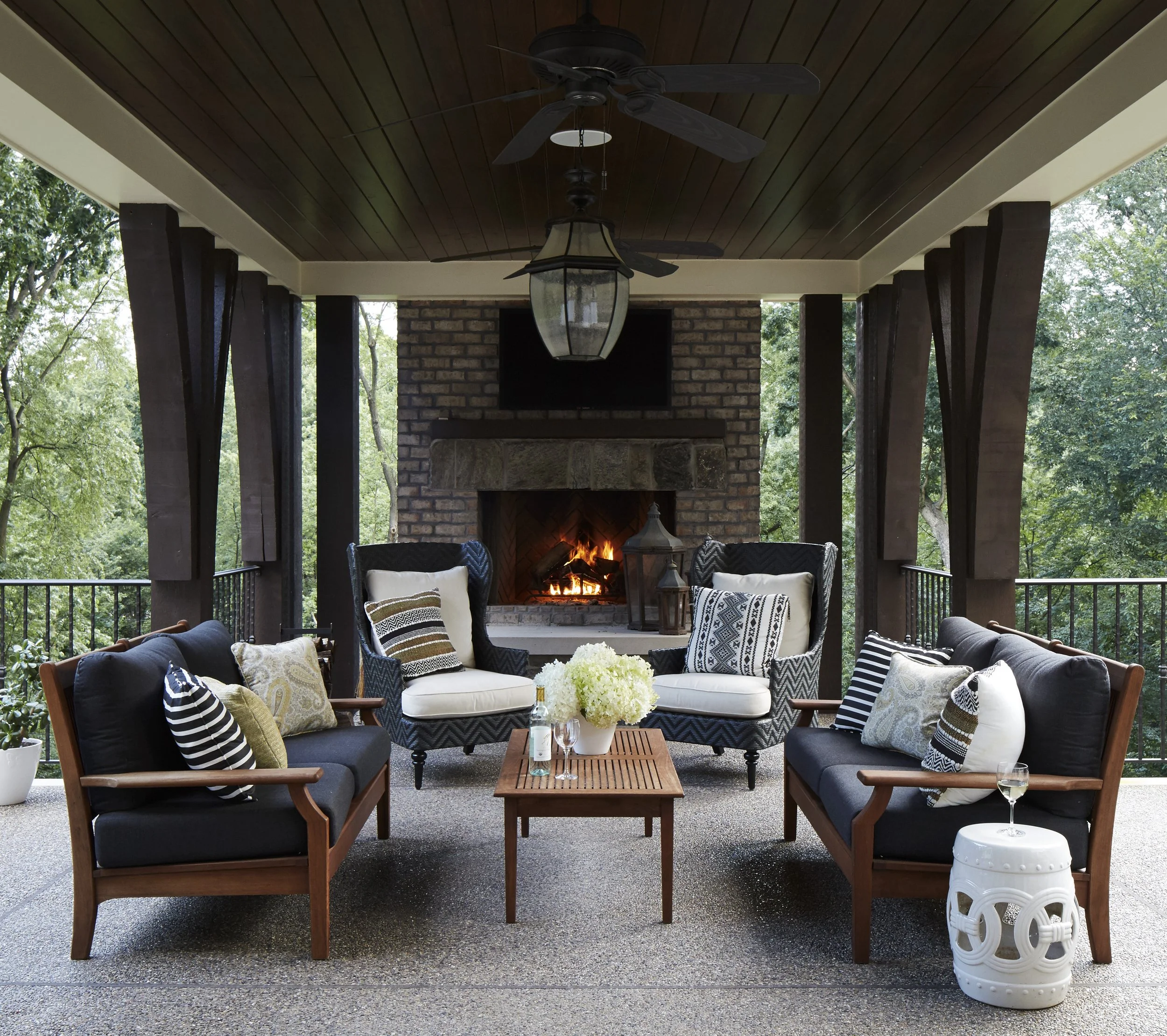 Covered outdoor patio with black wicker furniture and pillows, a wooden coffee table with a floral centerpiece, a brick fireplace with a fire burning, ceiling fans, and a view of green trees in the background.