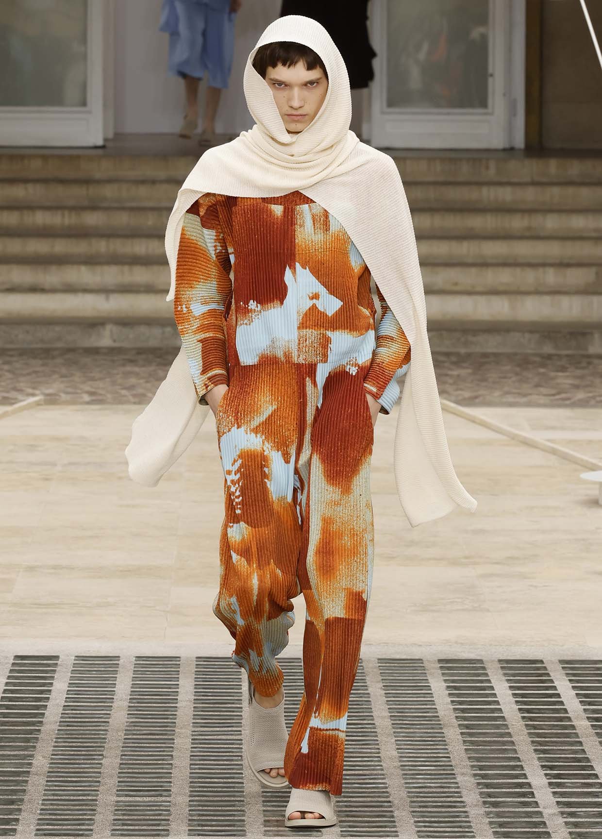 Model walking on runway wearing vibrant orange, white, and light blue tie-dye patterned outfit with a white hooded scarf.