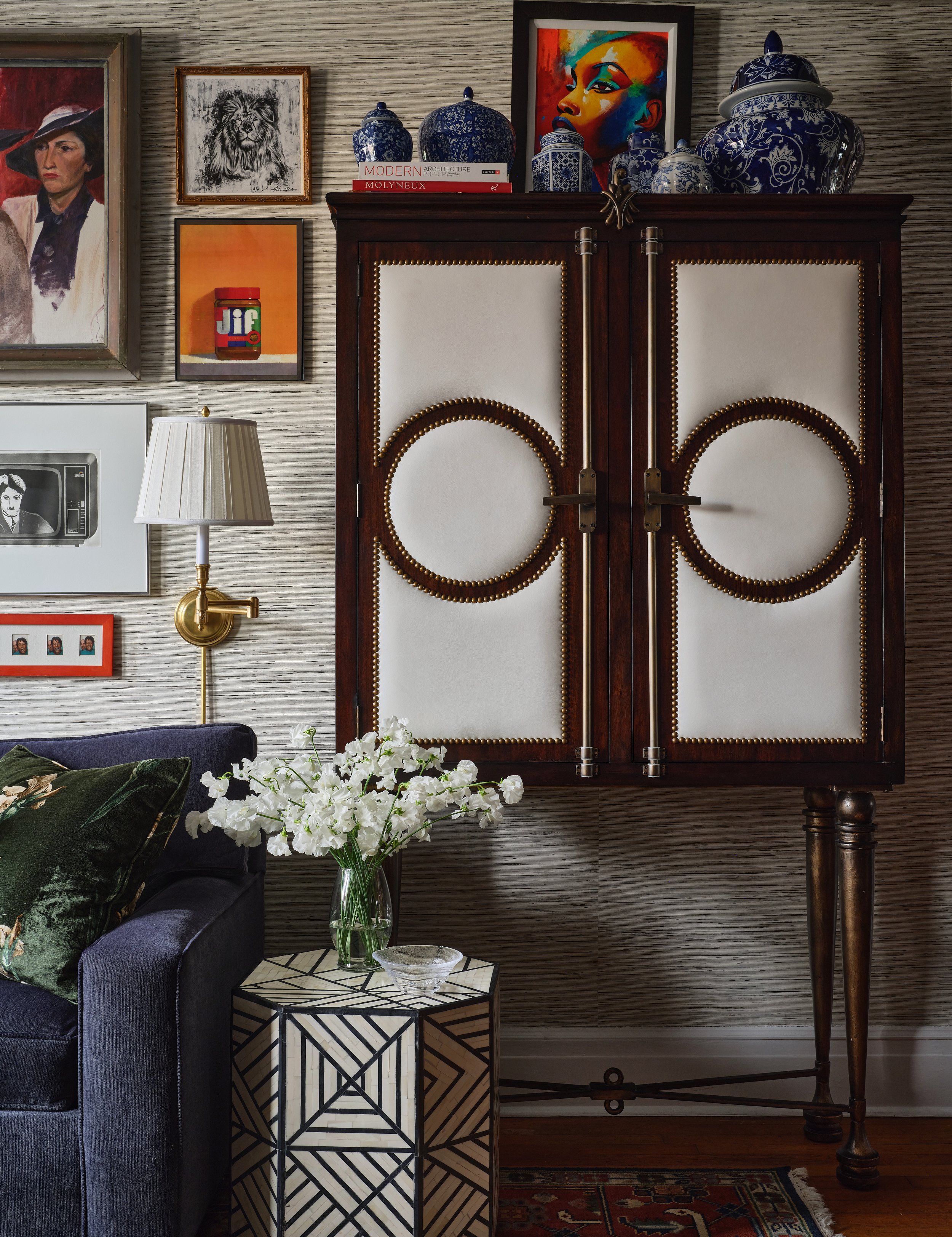 Interior living space featuring a dark wooden cabinet with ornate golden accents, decorated with blue and white porcelain jars. To the left, a gallery wall displays framed artwork including a painting of a woman in a hat, a monochrome lion sketch, a 