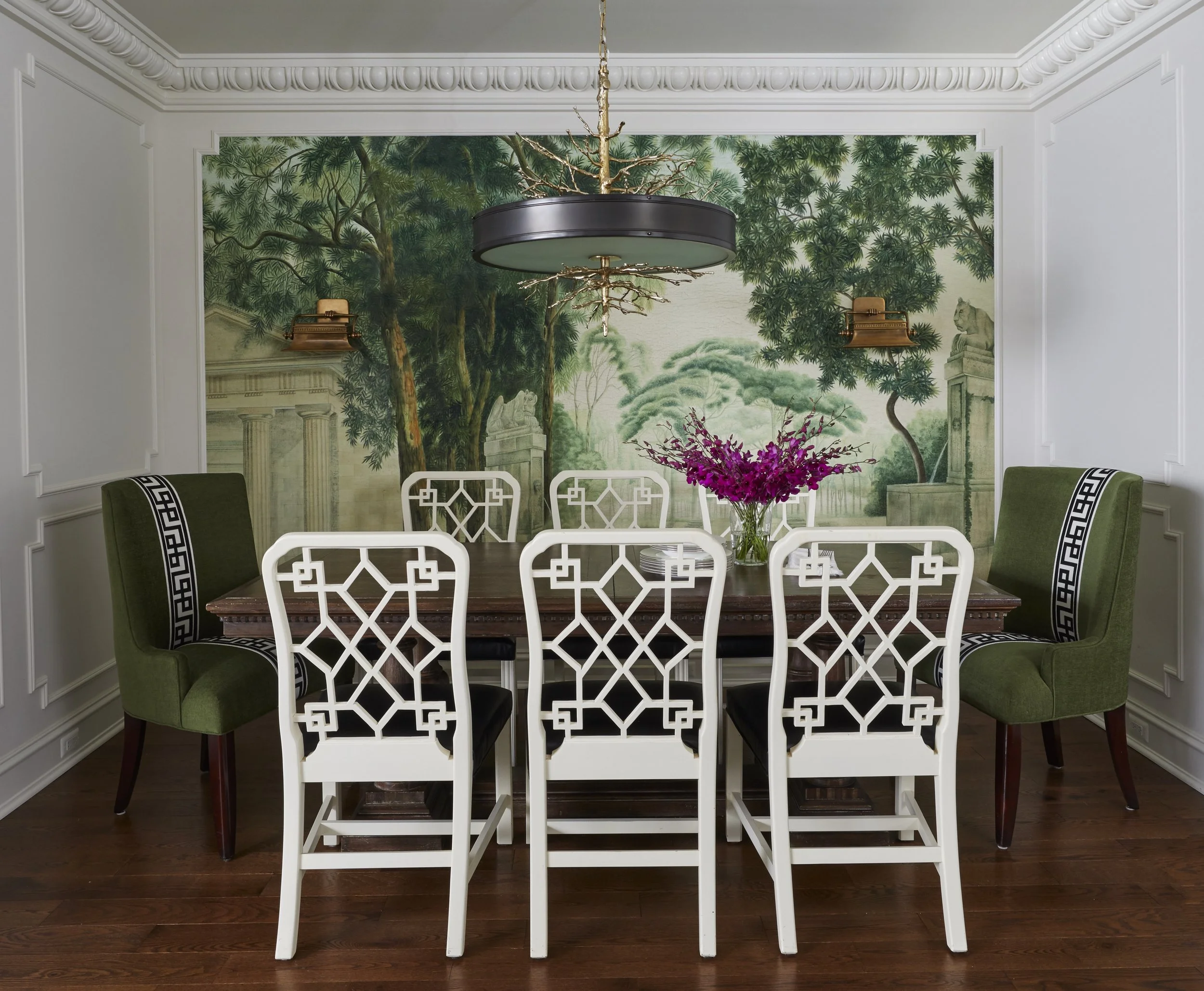 Dining room with a wooden table, white decorative chairs, green upholstered chairs, a floral centerpiece with pink flowers, a mural of trees and statues on the wall, and a modern chandelier hanging from the ceiling.