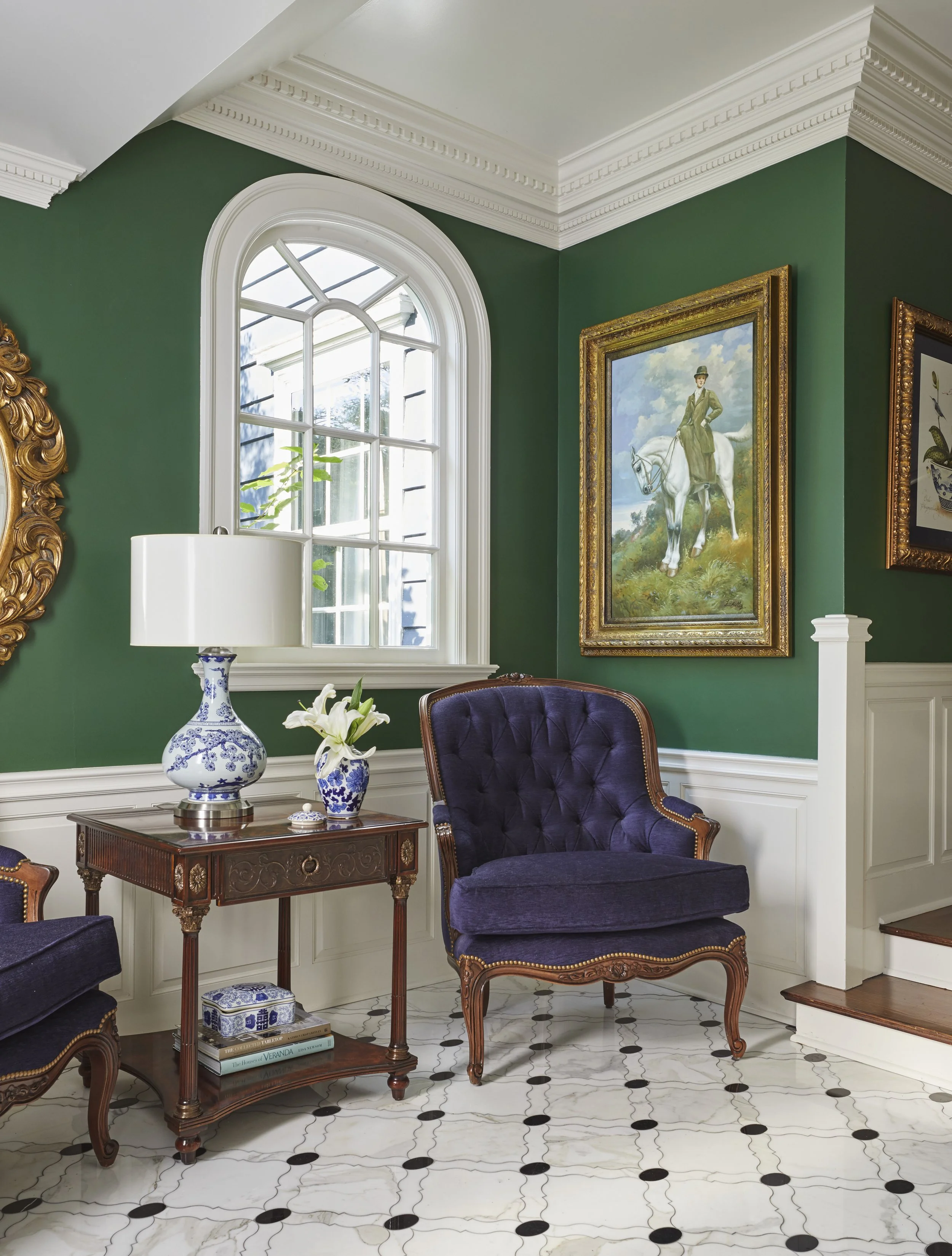 A corner of a room with green walls, decorative white crown molding and trim, a white arched window, and framed paintings. The furniture includes a purple upholstered armchair, a wooden side table with blue and white vases, and a white lamp. The floo