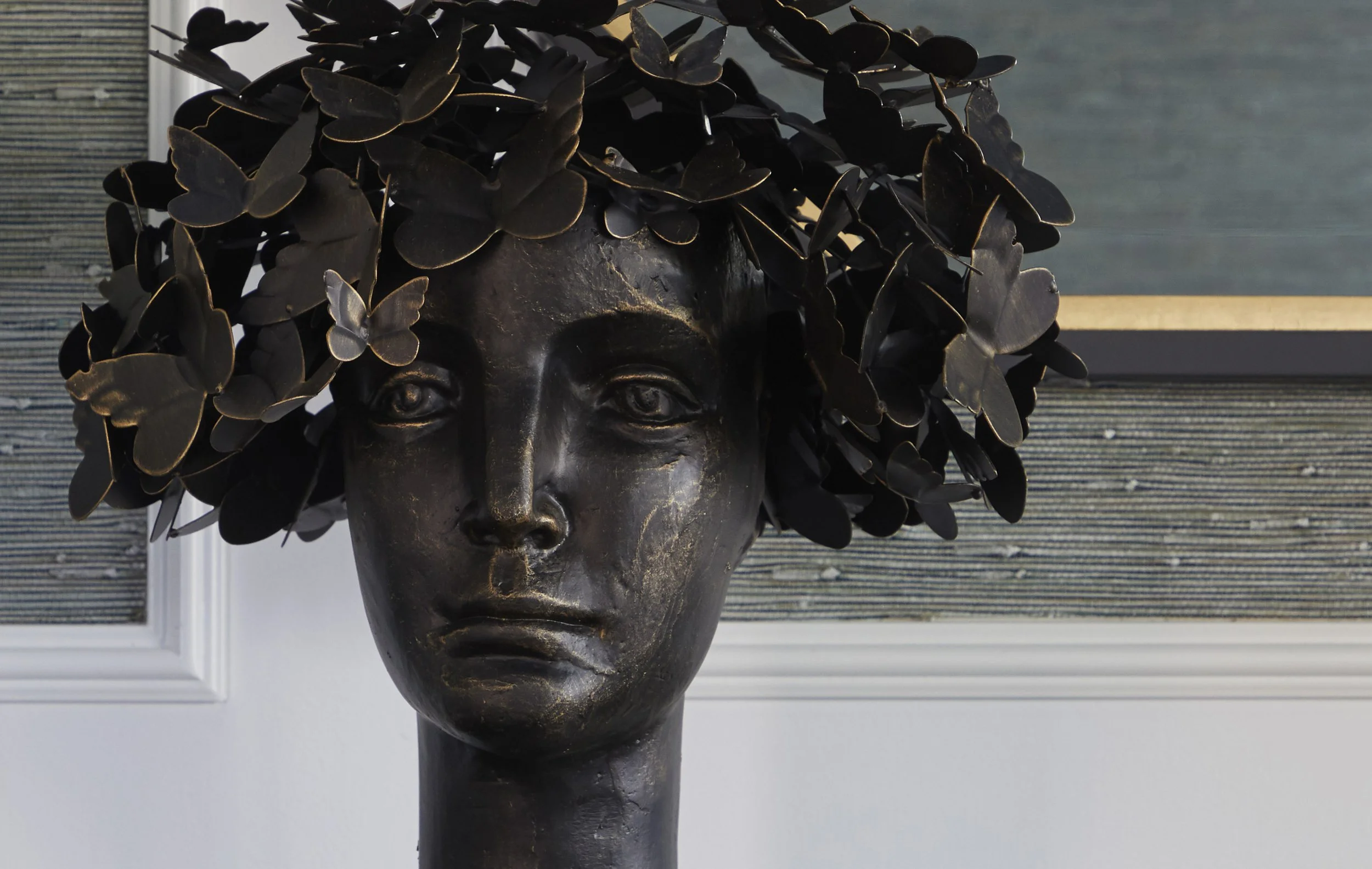 A dark-colored sculpture of a human face with closed eyes and worn features, adorned with numerous black butterflies as hair, set against a background of a painted wall with horizontal lines and a window.