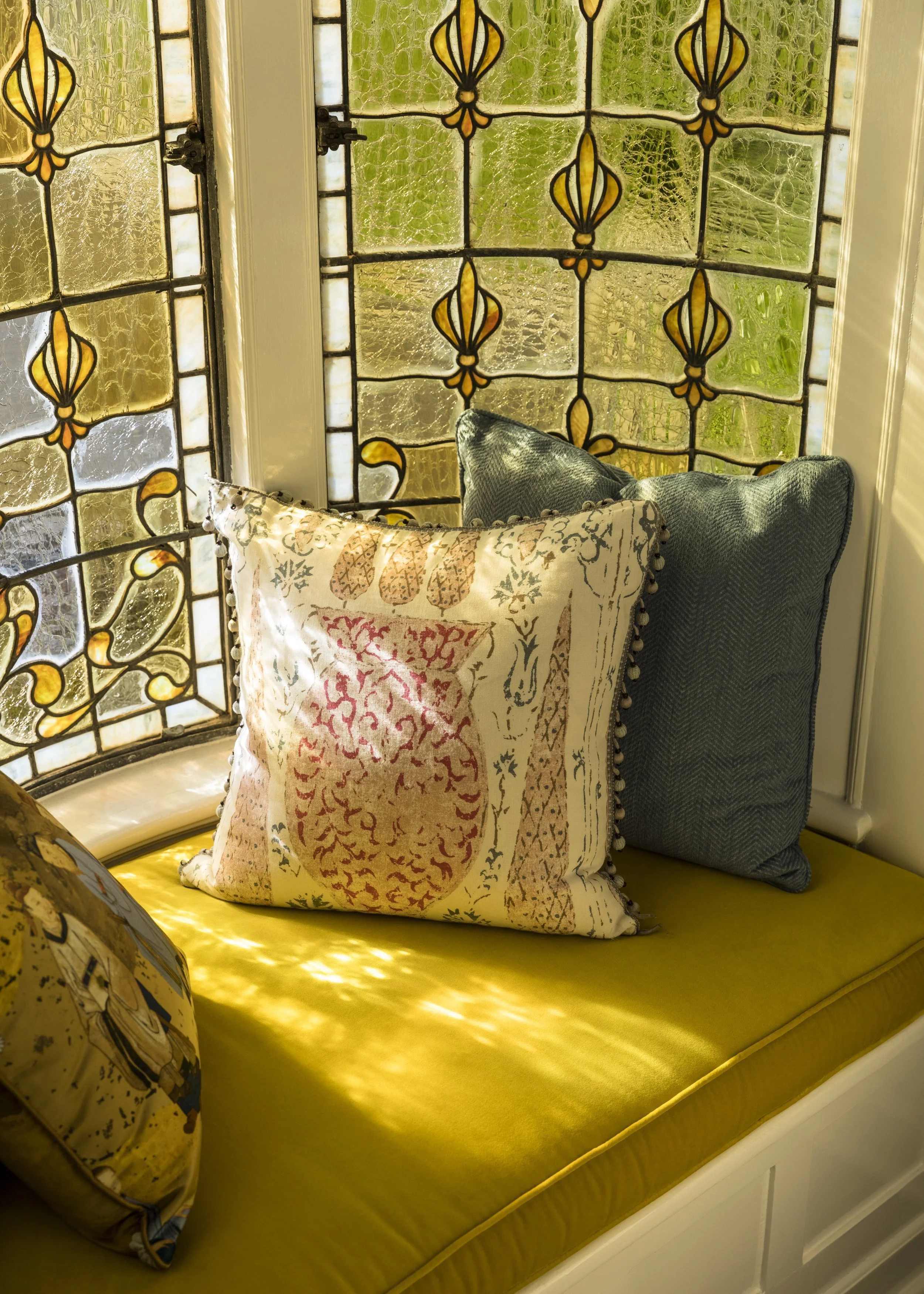 A window with stained glass featuring yellow and green floral patterns, with two decorative cushions on a yellow cushioned bench beneath it.