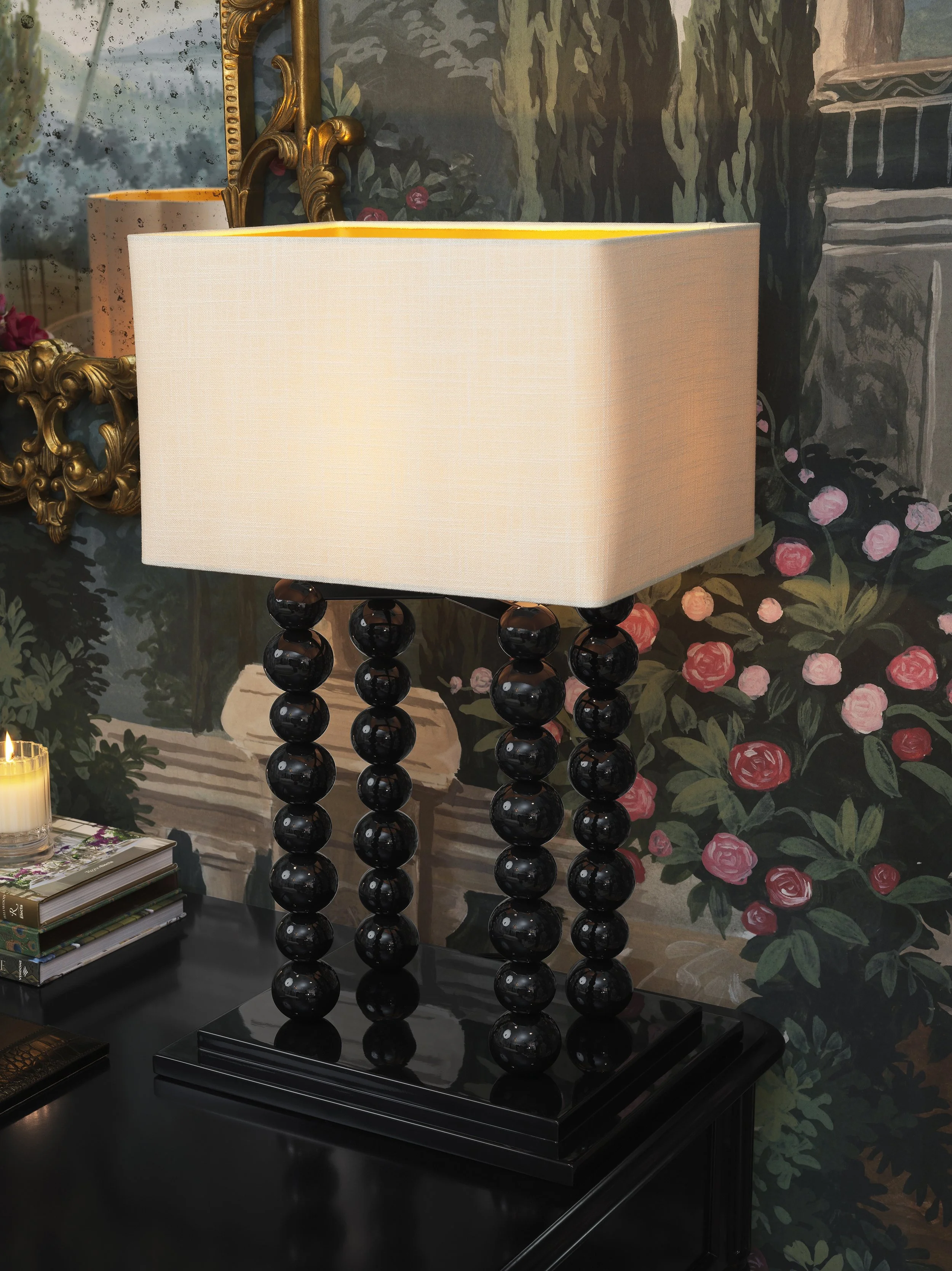 A black side table holds a lamp with a white rectangular shade with rounded edges, resting on a base made of glossy black spherical beads. Behind the table, a wallpaper features a floral and scenic design with bushes, flowers, a staircase, and a buil