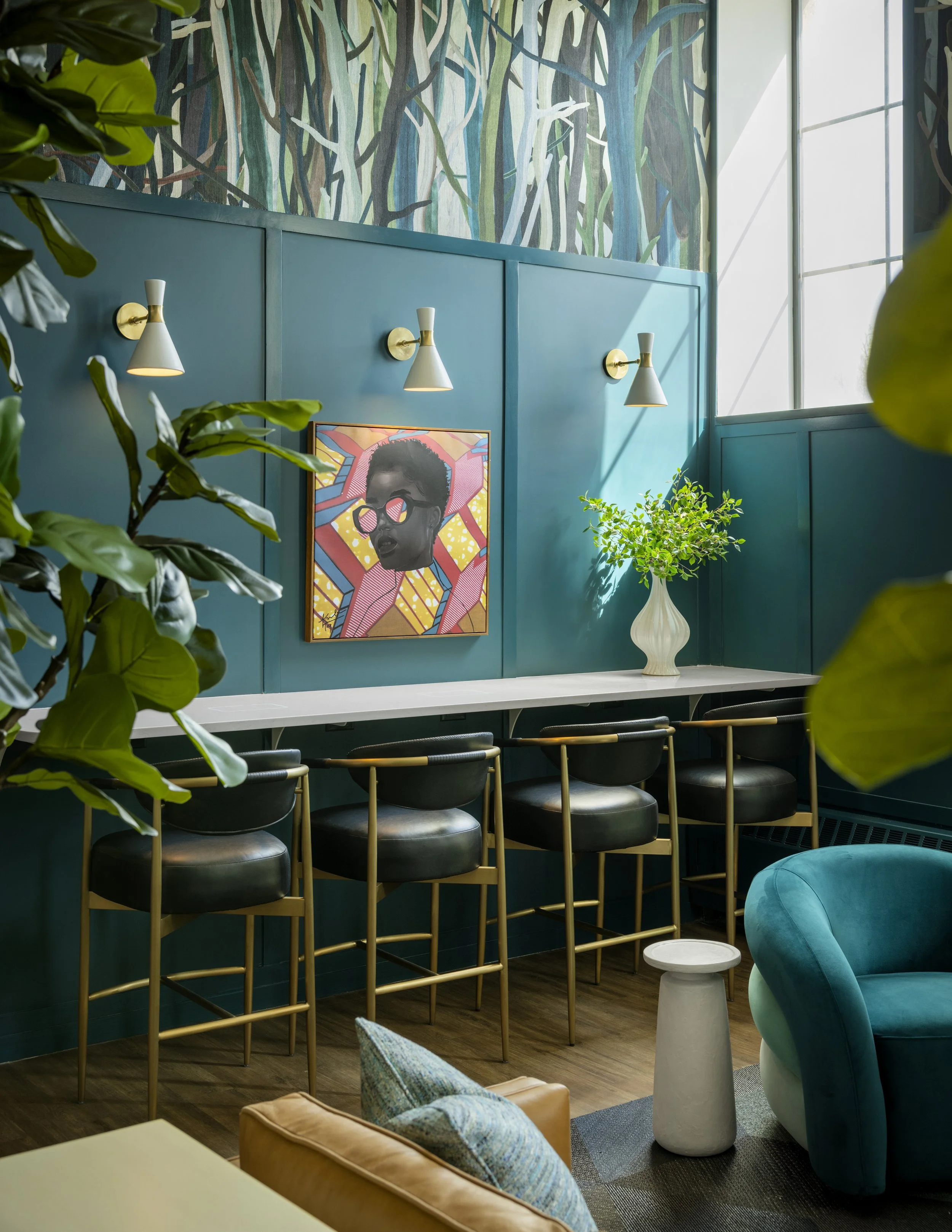 Interior of a modern room featuring a teal wall with three white wall-mounted lamps, a colorful portrait of a woman with sunglasses, green plants, and crisp natural light coming through large windows.