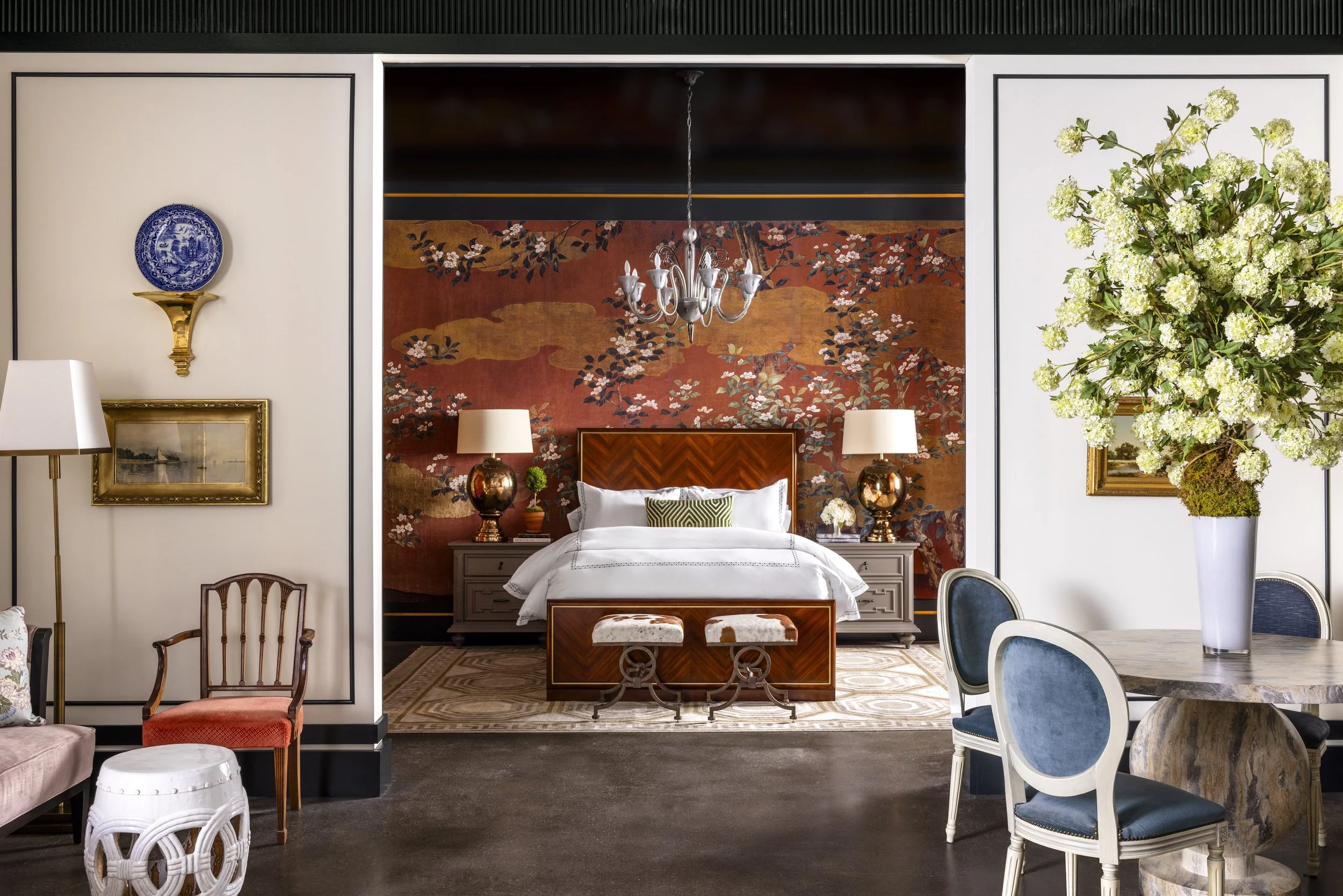 A luxurious bedroom with a patterned red-orange floral and nature wallpapered wall, central white bed with a padded headboard, flanked by two lamps on nightstands, and a chandelier hanging from the ceiling. The room is framed by white walls with blac