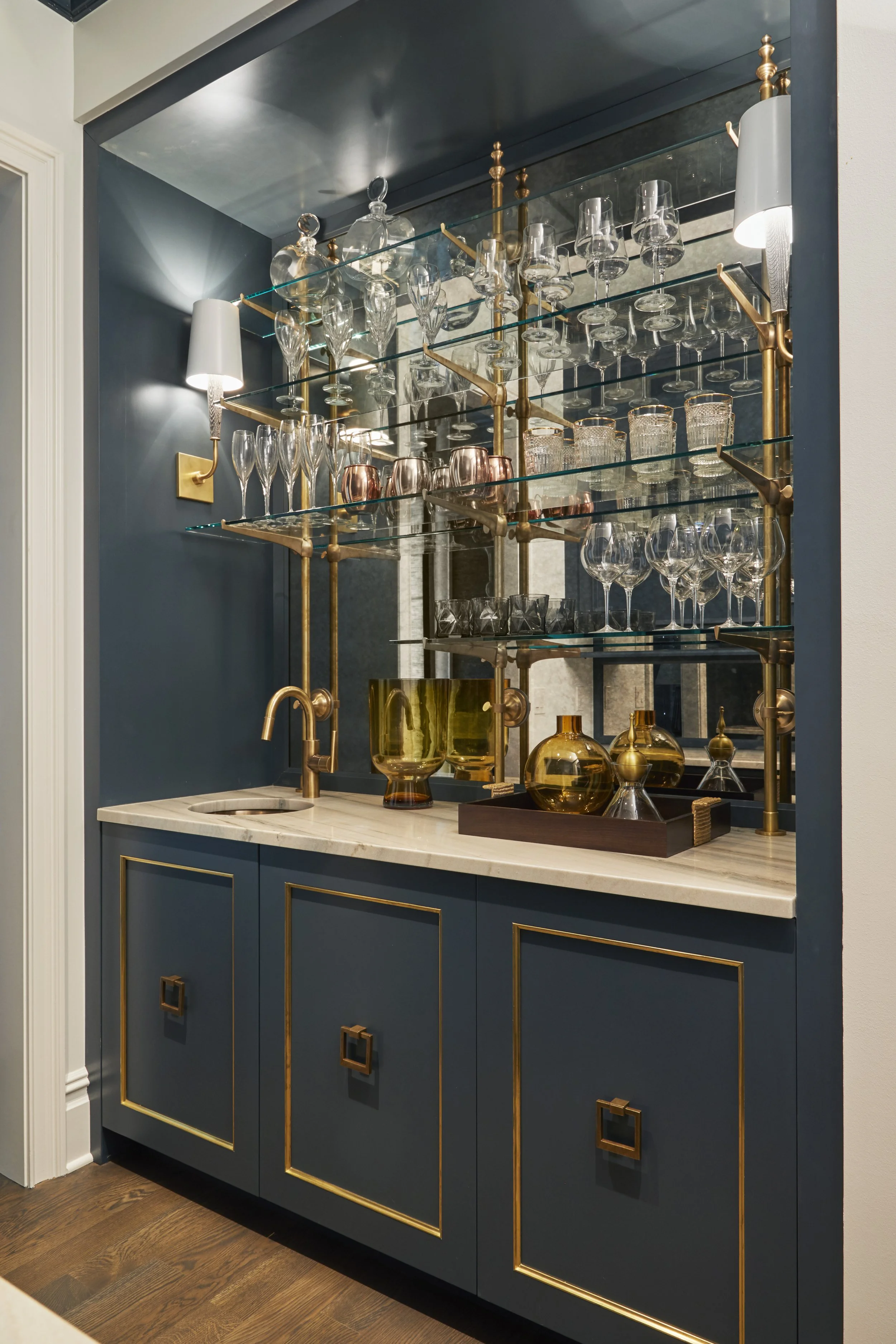 A bar cabinet with glass shelves holding various glassware and bottles, gold accents, and a marble countertop.