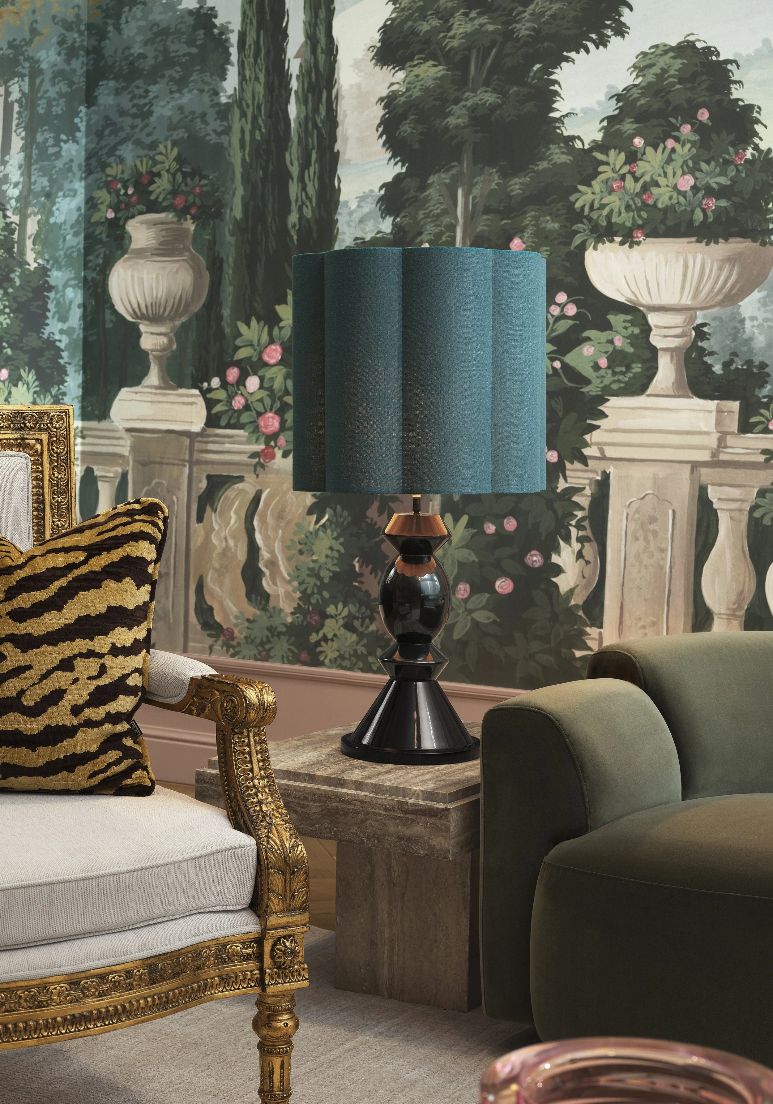 A room decorated with an ornate gold-trimmed white sofa, a green armchair, a marble side table with a modern black and copper table lamp, and a large mural of a garden with classical urns filled with pink roses and greenery.