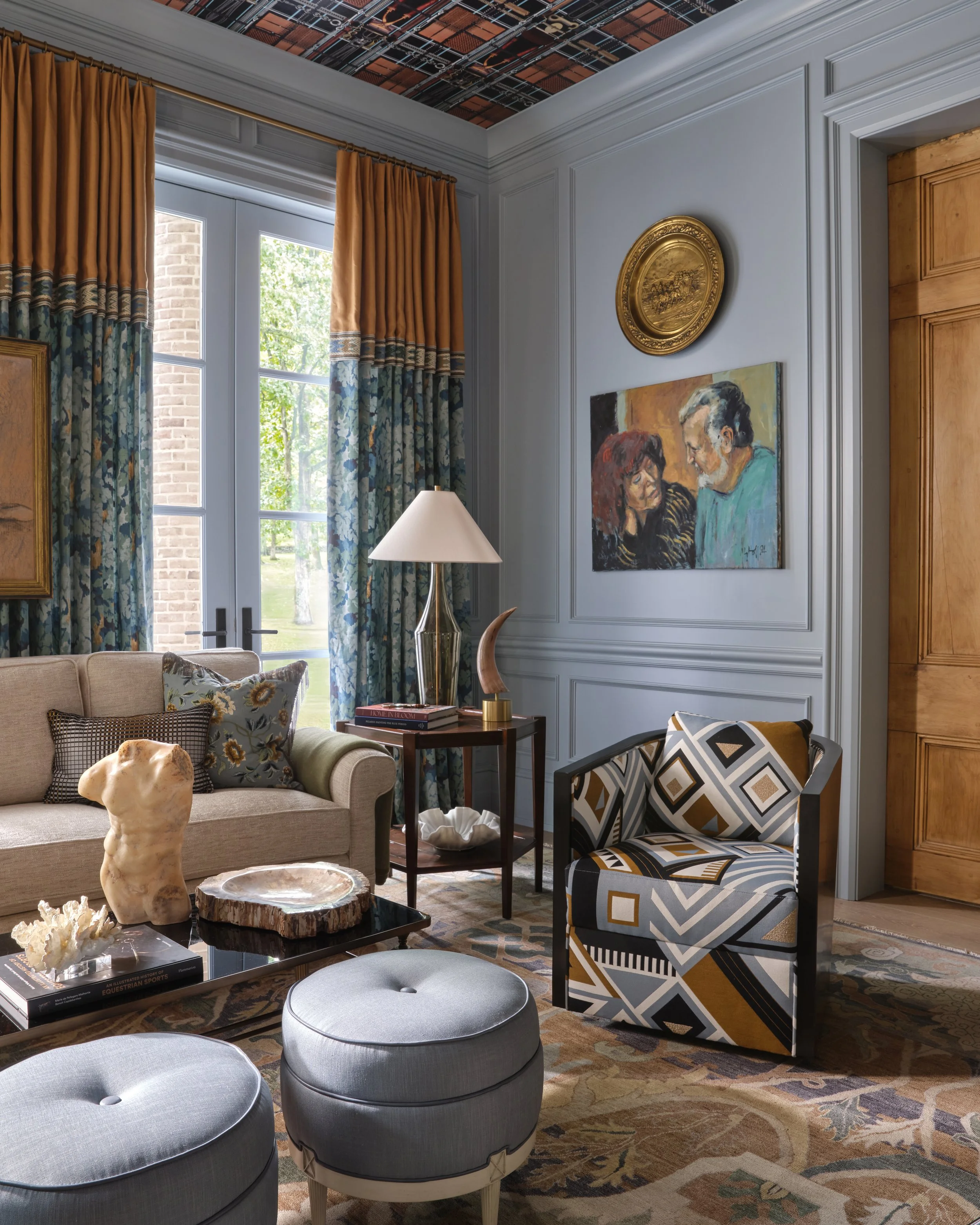 Living room with large window with orange and floral curtains, a beige sofa with pillows, a wooden side table with a lamp and books, a colorful patterned armchair, a black coffee table with decorative items, and wall art including an oval gold frame 