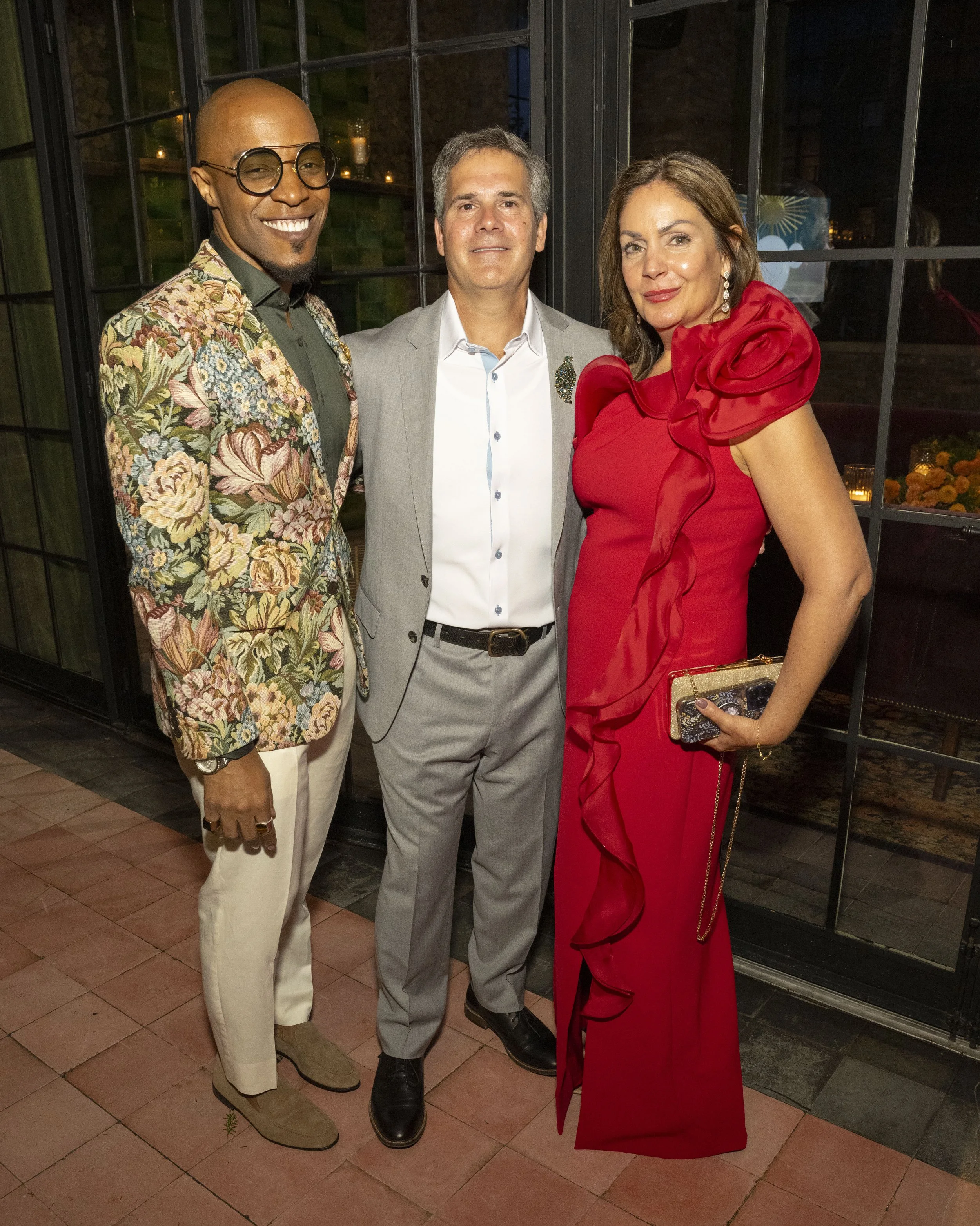 Three people dressed formally, standing inside near a large window with dark frames. The man on the left is wearing a floral jacket, light pants, and glasses, smiling confidently. The man in the middle is wearing a light grey suit, white shirt, and a decorative brooch, with a neutral expression. The woman on the right is wearing a red dress with ruffled sleeves and holding a clutch purse, slightly smiling, with earrings. The background suggests an evening event with dim lighting and decorative candles.