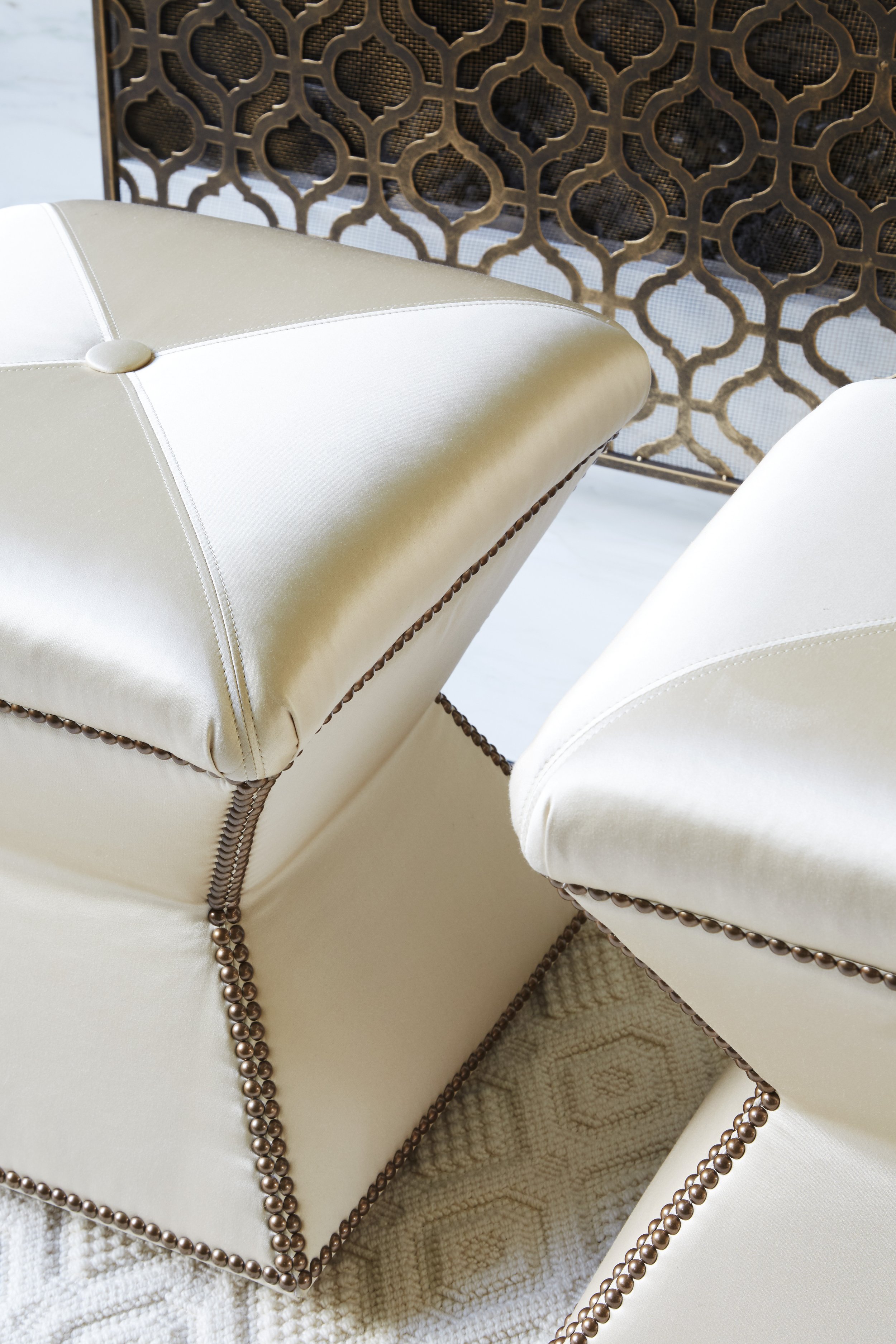 Close-up of cream-colored upholstered furniture with decorative bronze nailhead trim, set on a textured beige rug, with a wooden lattice decorative screen in the background.