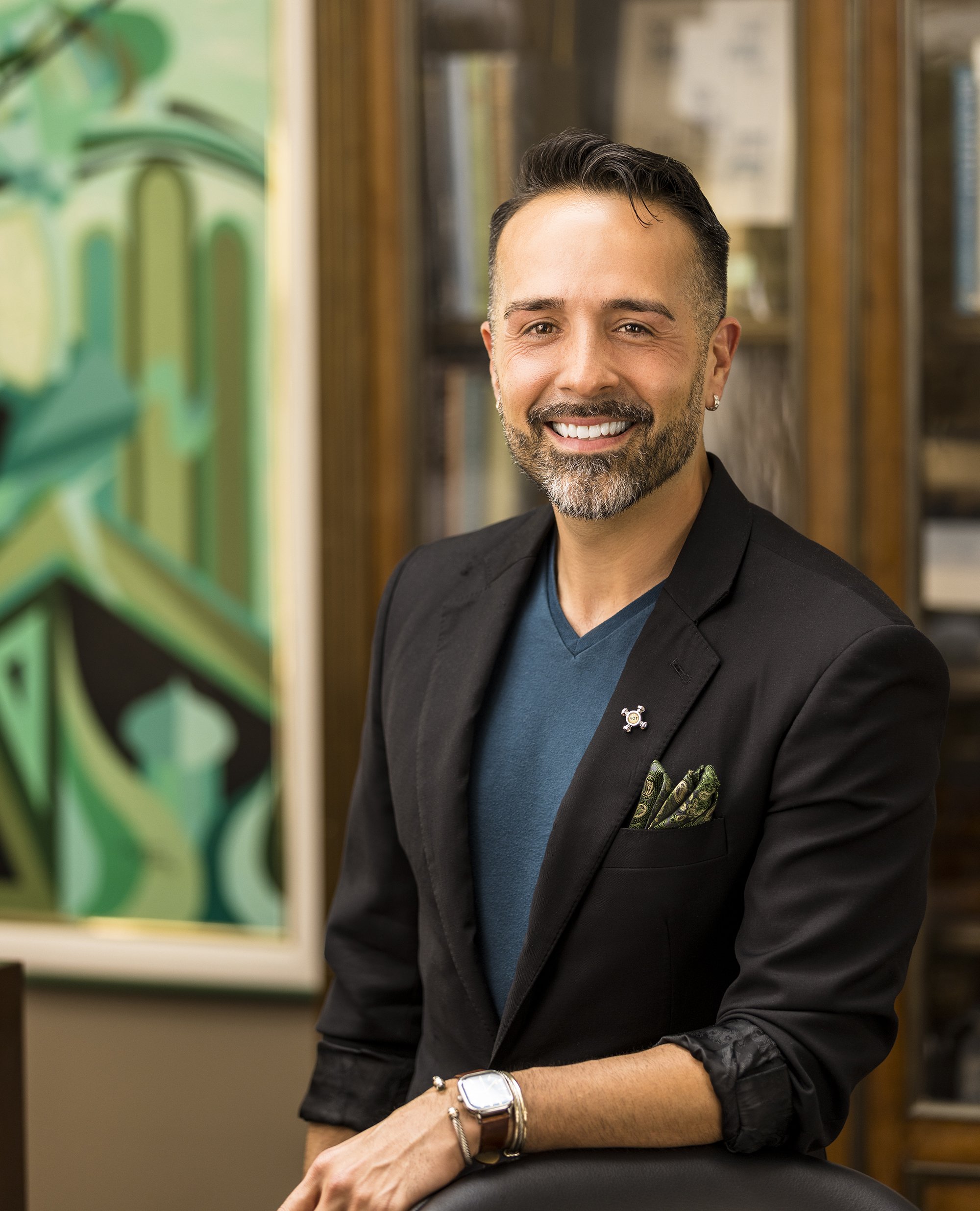 A man with short dark hair, a beard, and earrings, smiling, dressed in a black blazer over a blue shirt, wearing a silver watch and bracelet, sitting in front of a bookshelf with framed documents and a green abstract painting.