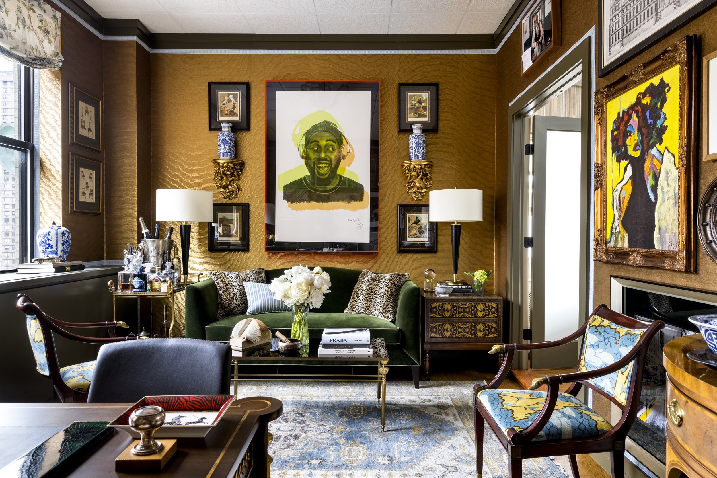 Interior of a decorated living room with gold textured walls, framed artwork, and eclectic furniture including a green velvet sofa with patterned pillows, a wooden table, and chairs with floral and animal patterns. The room has a large window with fl