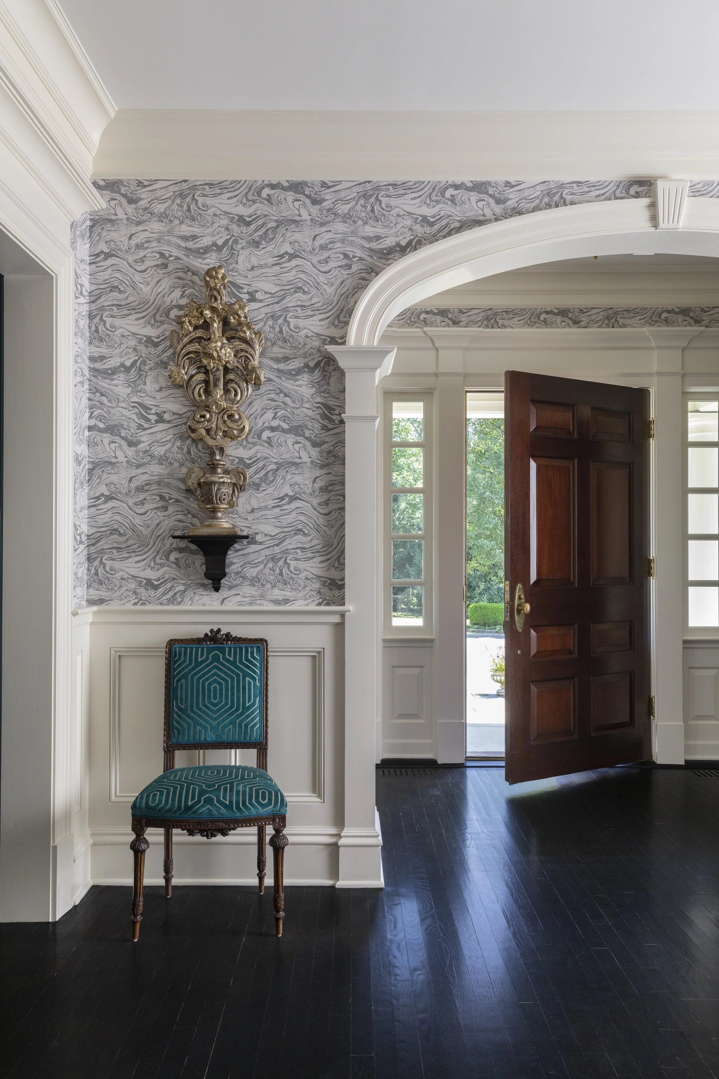 Interior view of a house entryway with a decorative wall, a wooden front door ajar, a blue patterned chair against the wall, and a wall sconce on the left. Bright sunlight outside through a window.