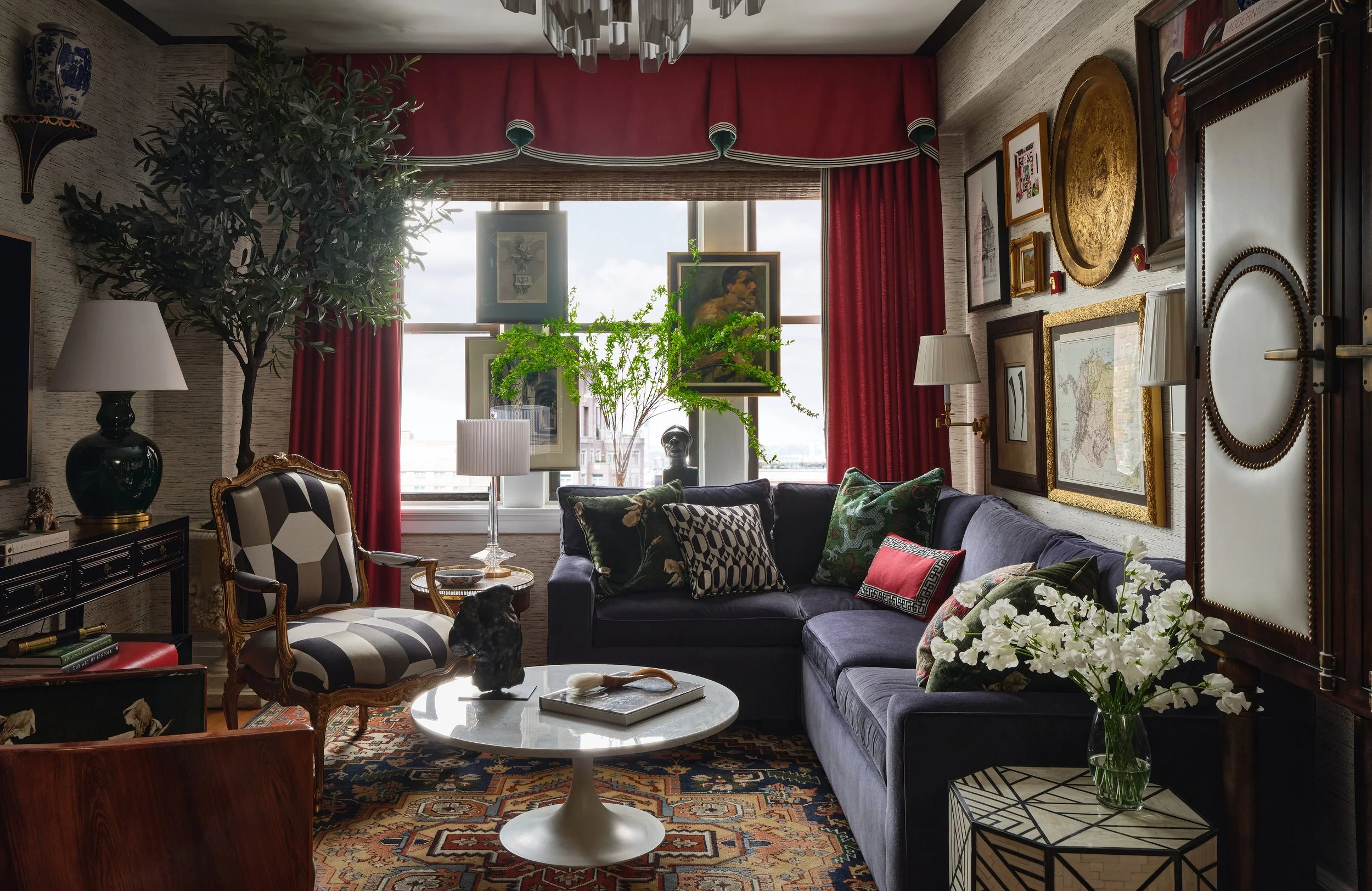 Living room with a large window, various paintings, and a mix of modern and vintage furniture, including a black sofa, a white marble-topped coffee table, and patterned pillows, decorated with plants and lamps.