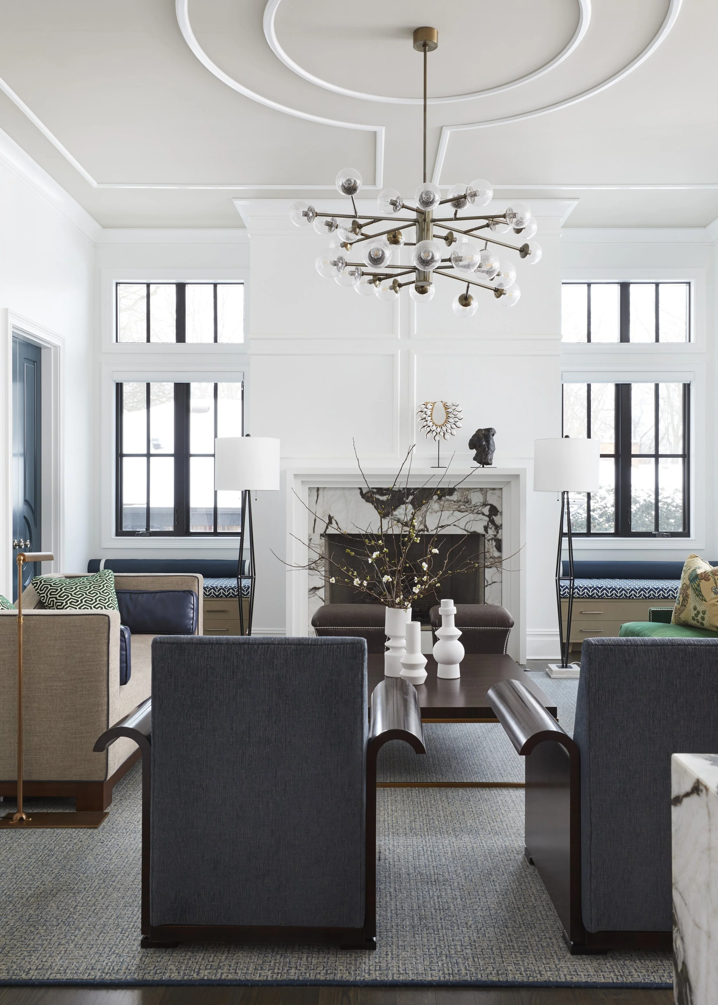 A modern living room with a large chandelier hanging from a ceiling with decorative molding. The room features a fireplace with a marble surround, two large windows flanking the fireplace, and a mix of seating including armchairs, benches, and a sofa