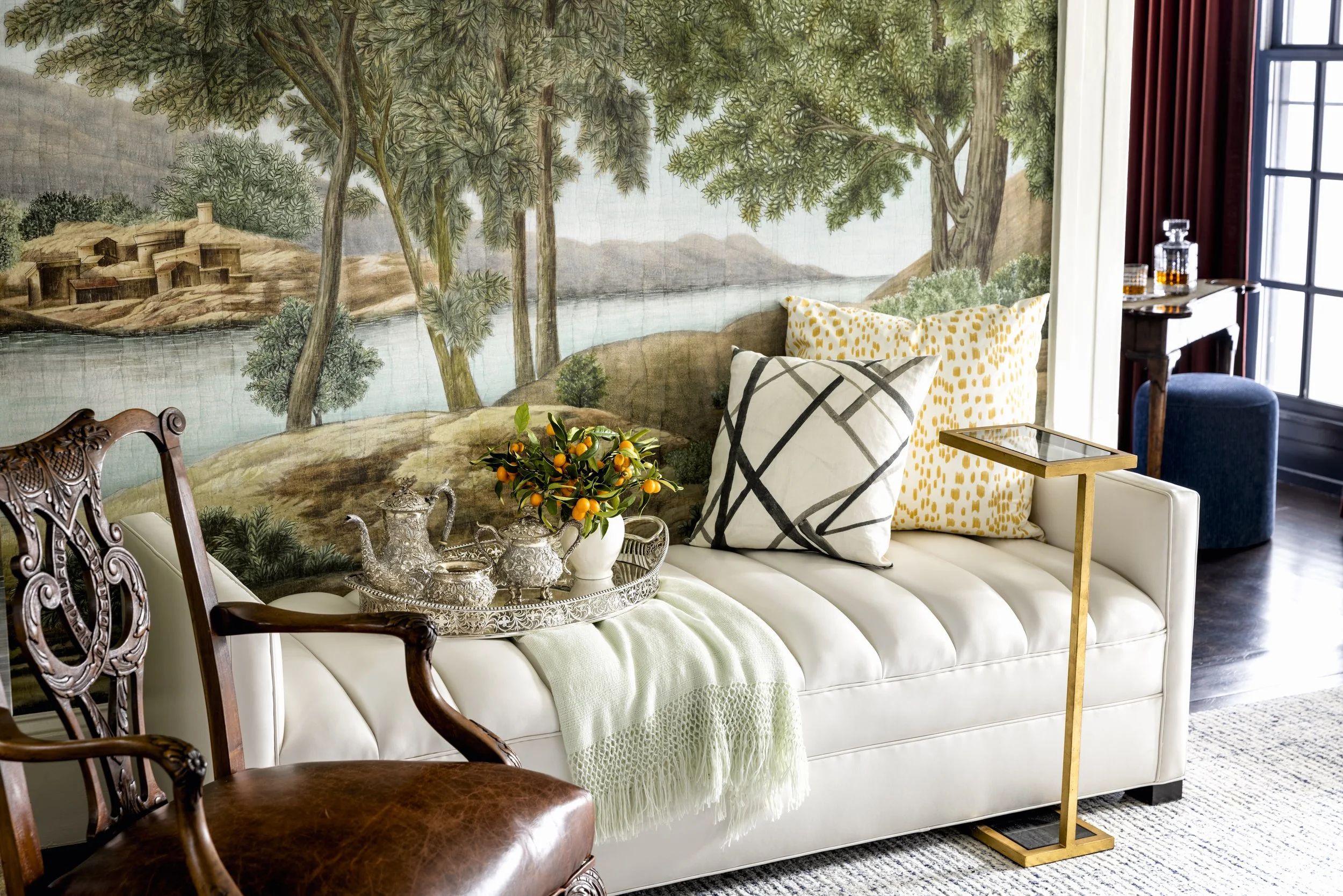 A living room with a white sofa, decorated with black and white pillows, and a small gold side table. Behind the sofa, there is a mural of trees, water, and hills painted on the wall. In front of the sofa, there is a silver tray with decorative items