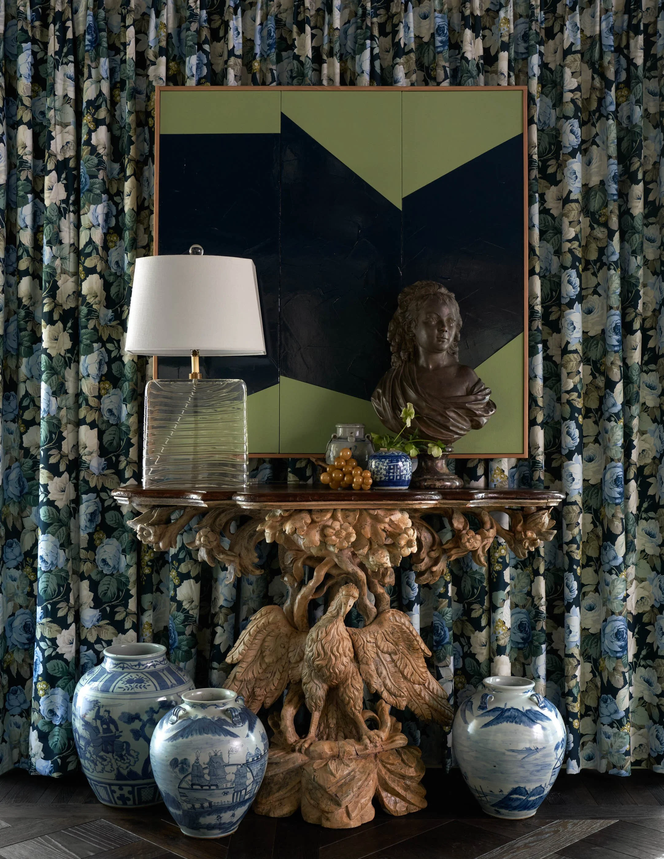 Decorative arrangement with a wooden carved table, blue and white porcelain vases, a bust sculpture, a table lamp, and a backdrop of floral patterned curtains.