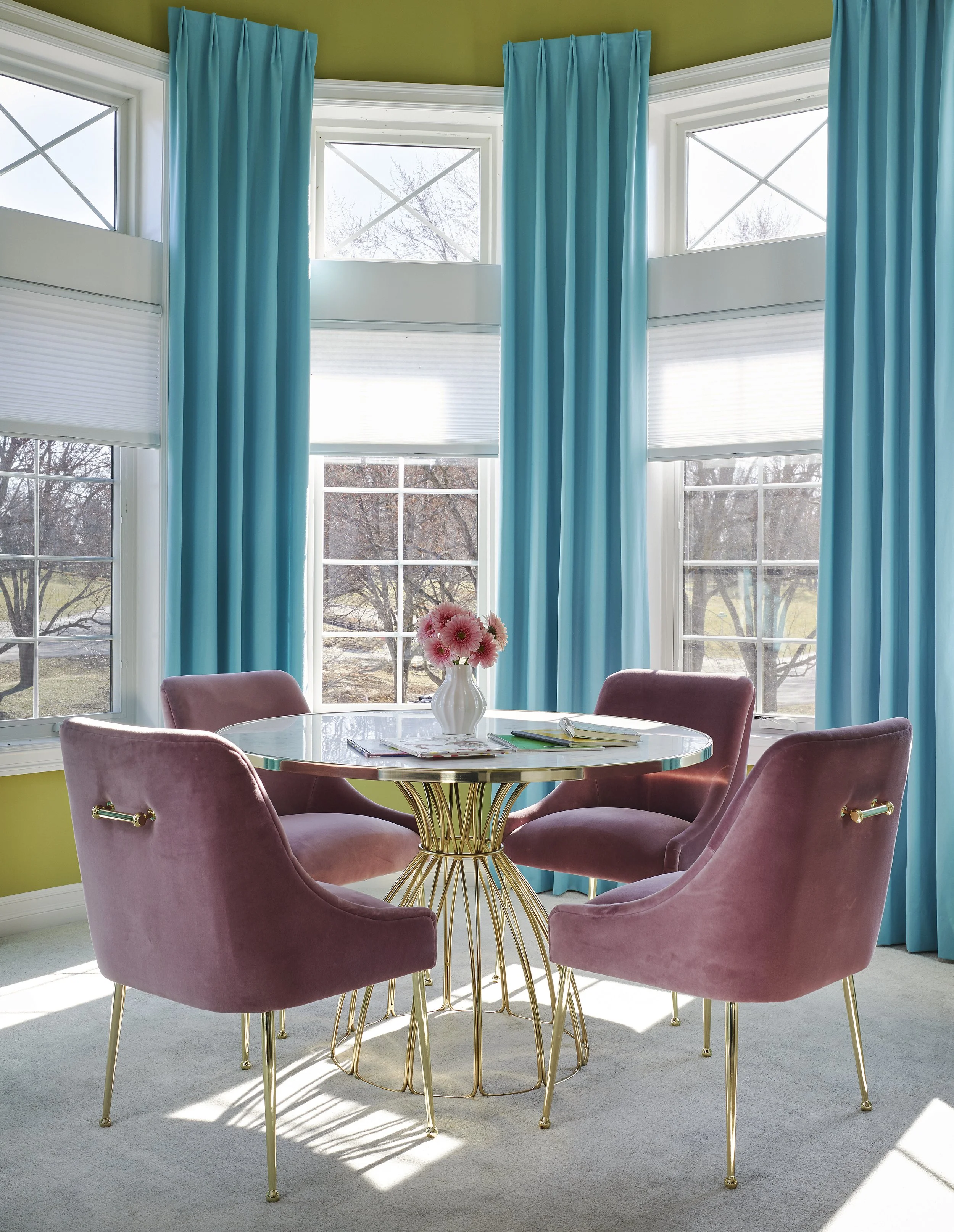 A dining area with a round glass table and four pink velvet chairs with gold legs, set near large windows with blue curtains and a vase of pink flowers on the table.