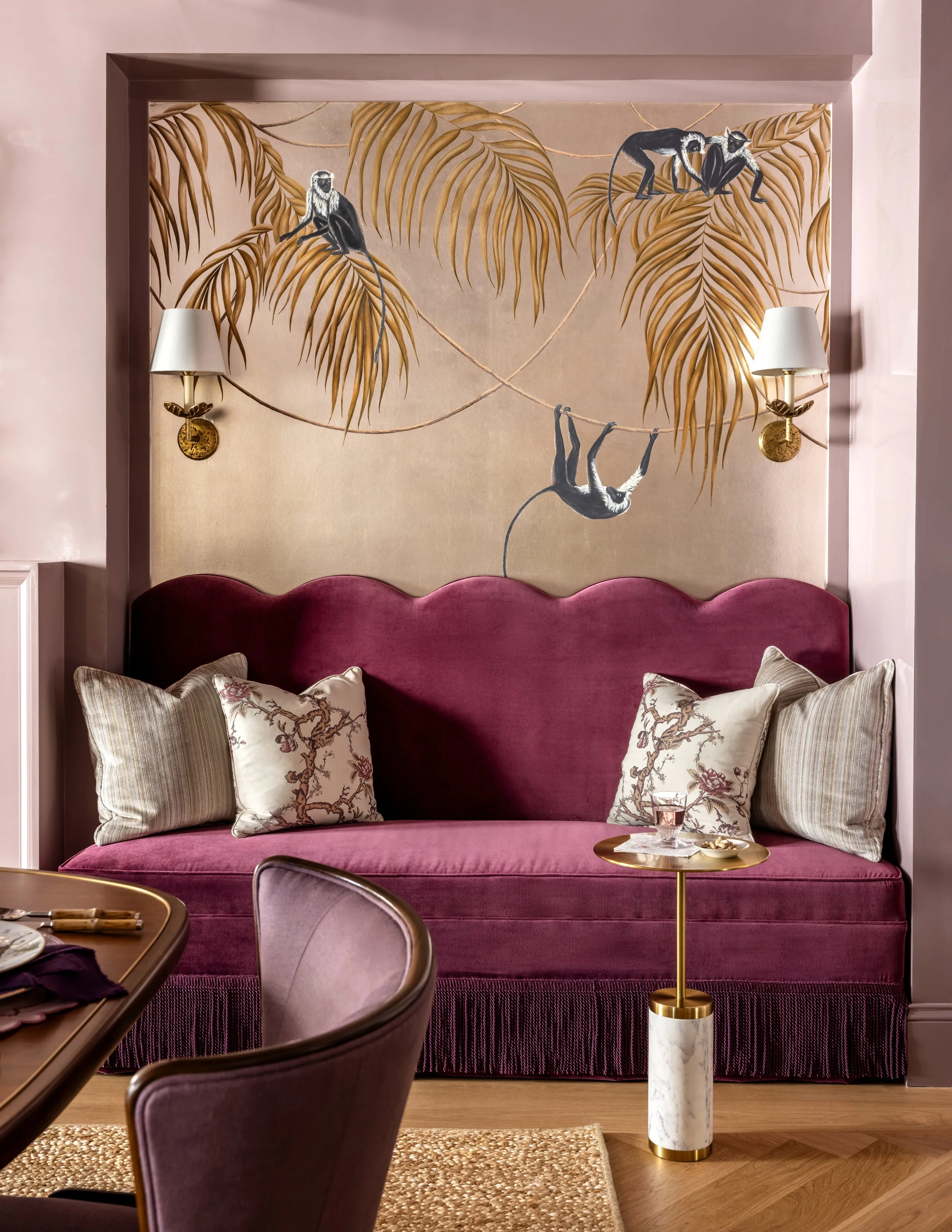 A pink upholstered seating area with decorative pillows, a pink wall, a gold and white marble side table with a glass of water and snack, and a wall mural of monkeys climbing on palm leaves with pink walls and gold wall-mounted lamps.