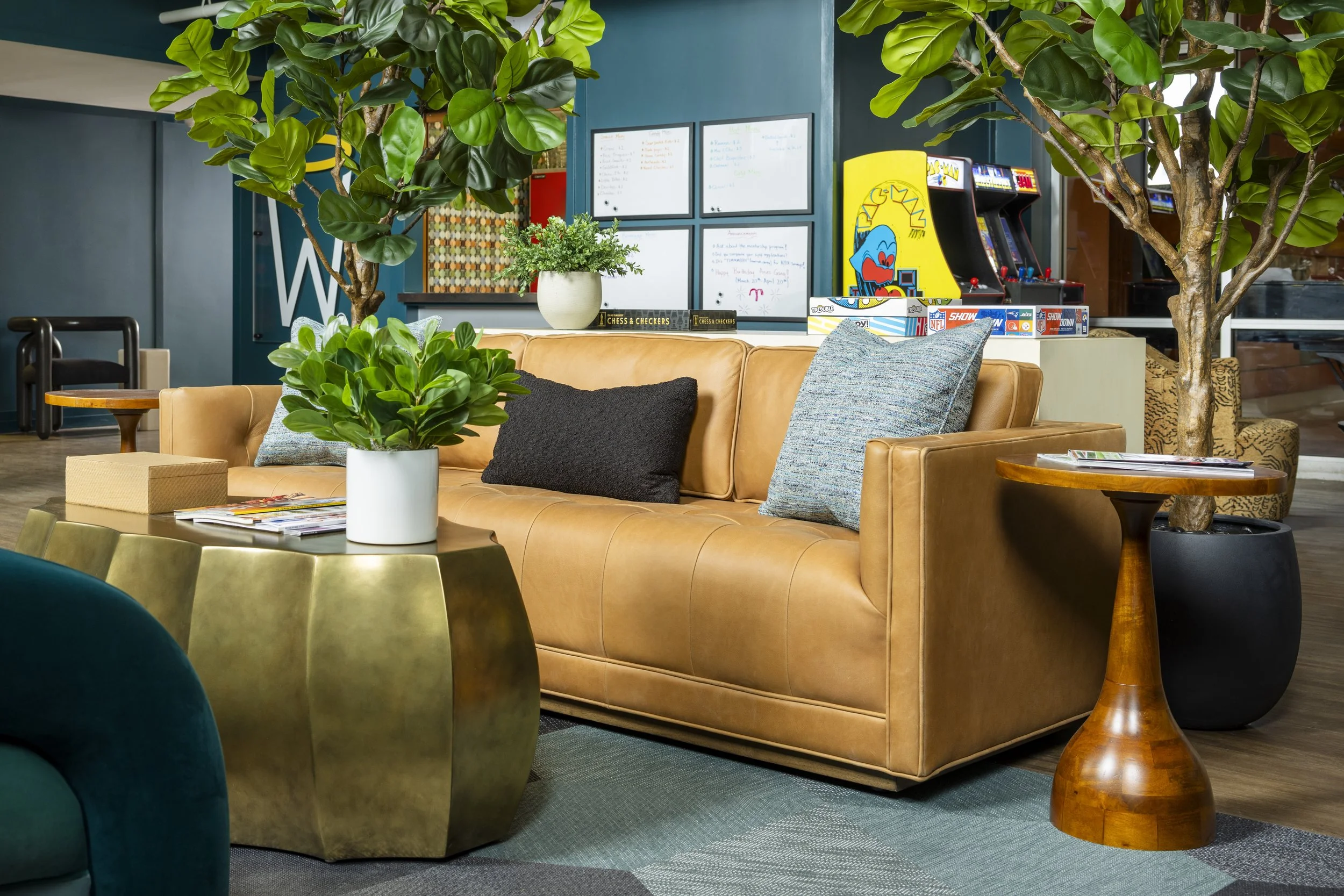 A cozy lounge area with a tan leather sofa adorned with gray and black pillows, surrounded by green indoor plants, a wooden side table, a puzzle game, and whiteboards on the wall in the background.