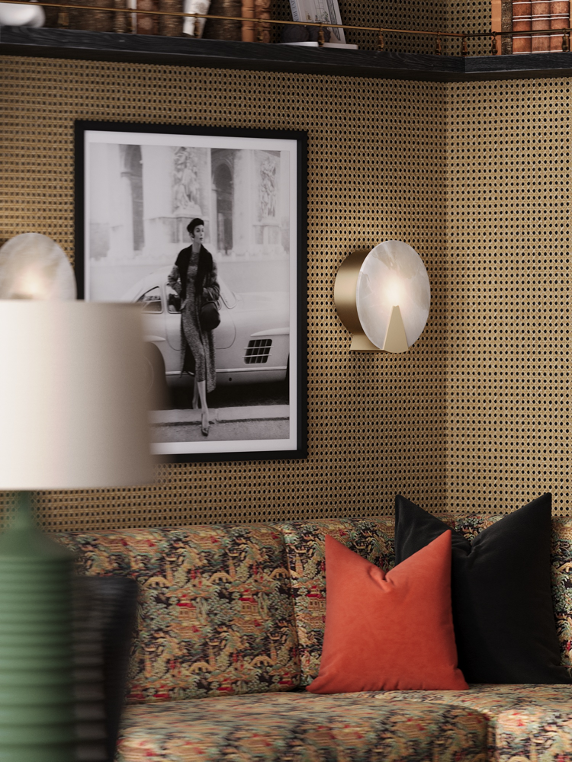 Living room with wallpapered wall, framed black and white photo of a woman near a vintage car, modern wall light fixtures, patterned sofa with red and black pillows, and a blurred lamp in the foreground.