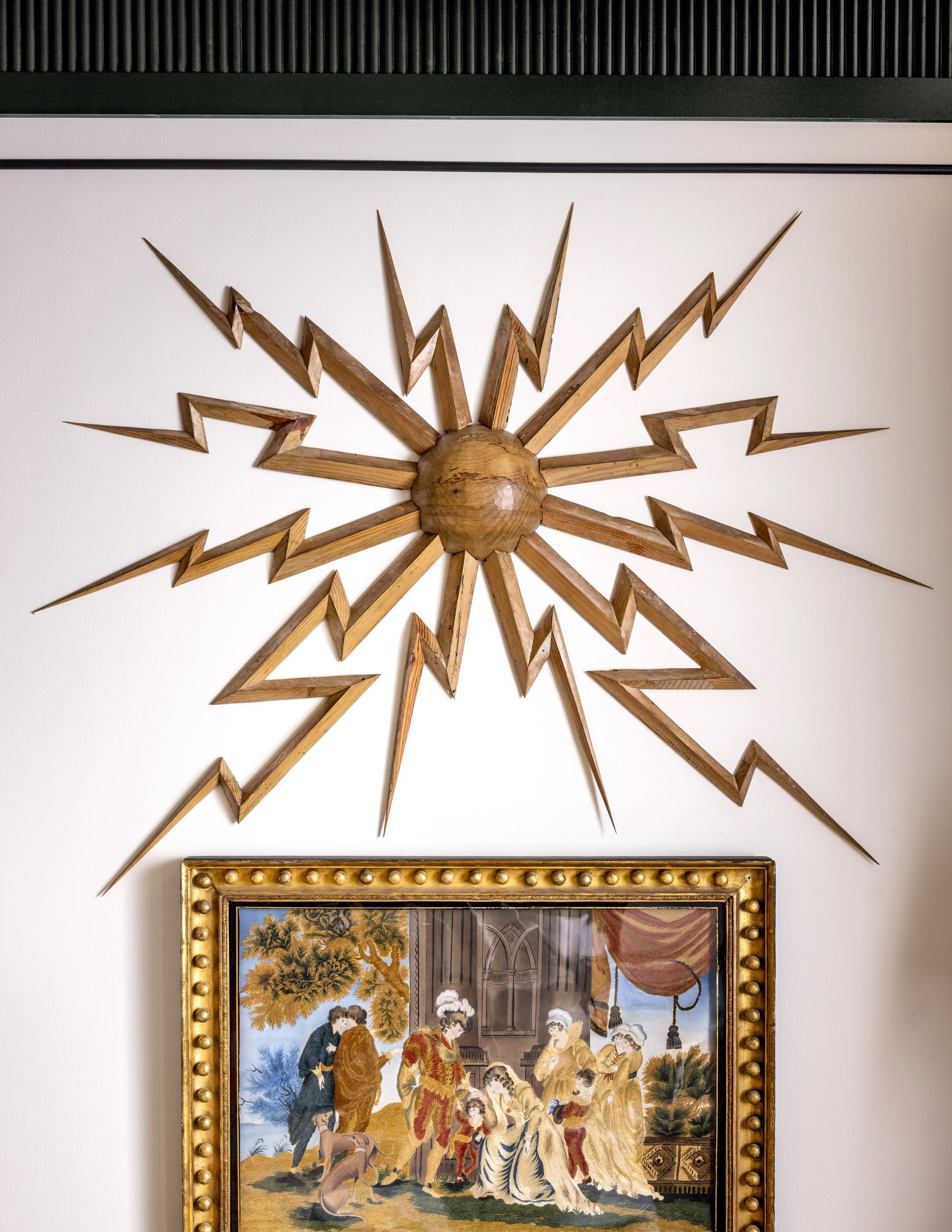 A decorative wooden sunburst wall sculpture mounted above a framed painting in a room. The painting depicts a scene with several people and animals, framed with a gold border featuring round embellishments.