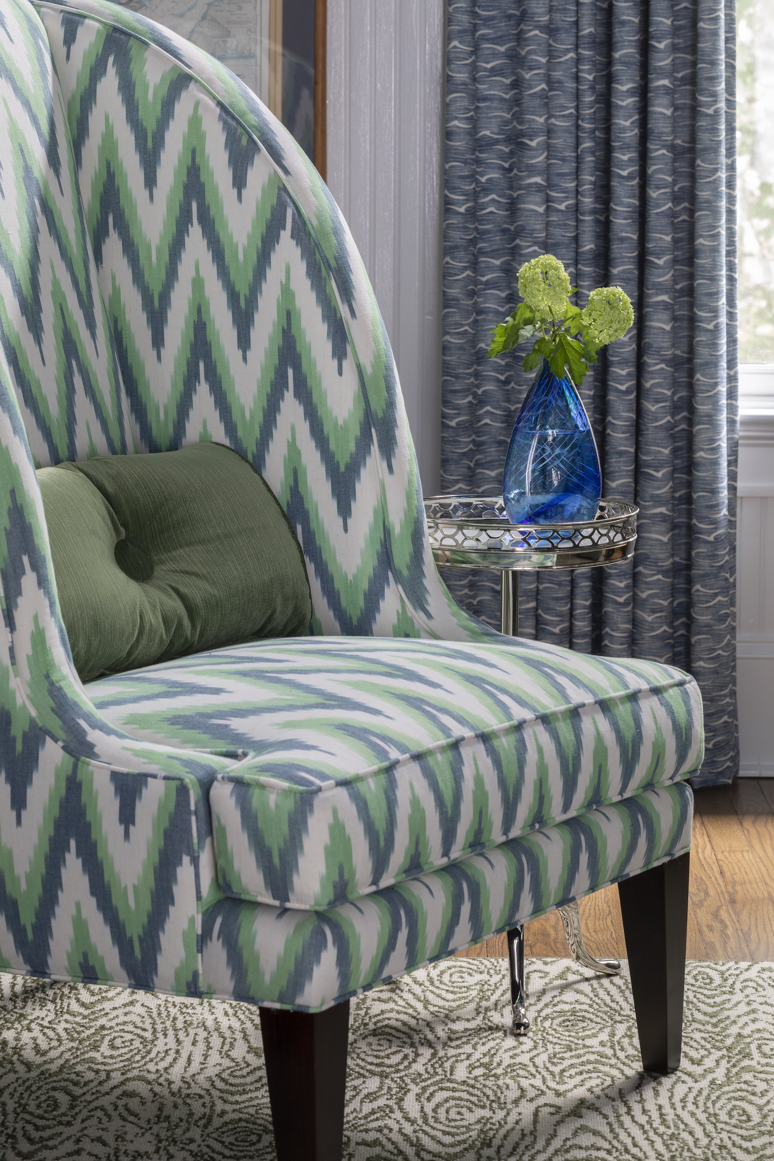 A room with a bed featuring a patterned headboard and mattress in shades of green, blue, and white. A green pillow rests on the mattress. Beside the bed, there is a round, silver side table with a blue glass vase containing green hydrangea flowers. B