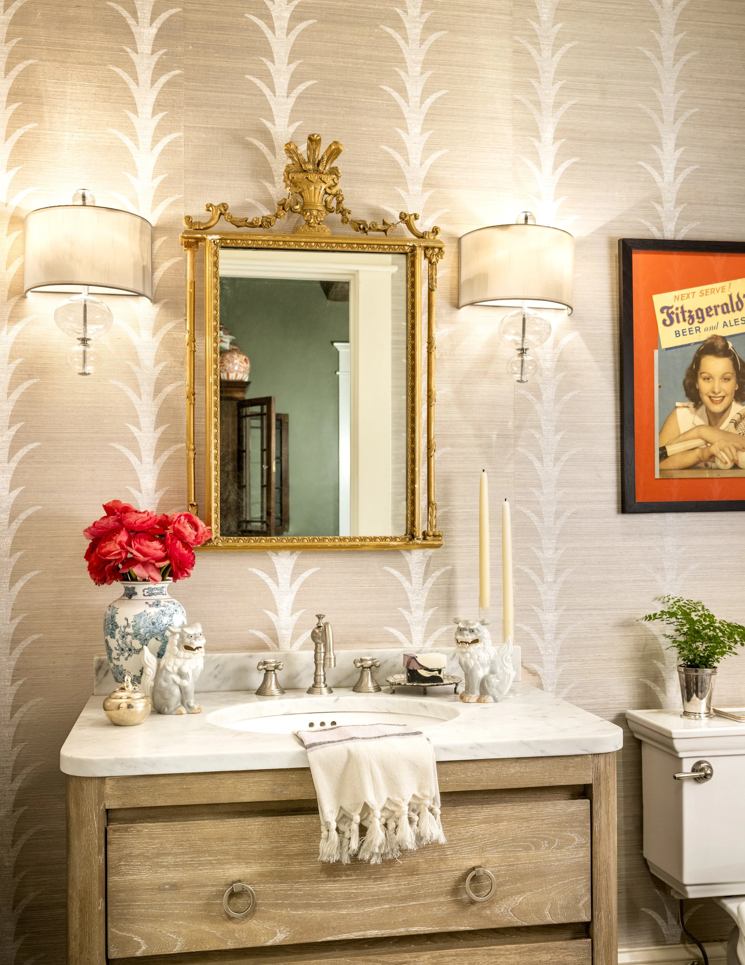 Bathroom vanity with a marble countertop, gold framed mirror, two ceramic lion figurines, a vase of pink flowers, a box of soap, and two candle holders with white candles. On the wall are two modern wall sconces and a framed vintage poster of a woman