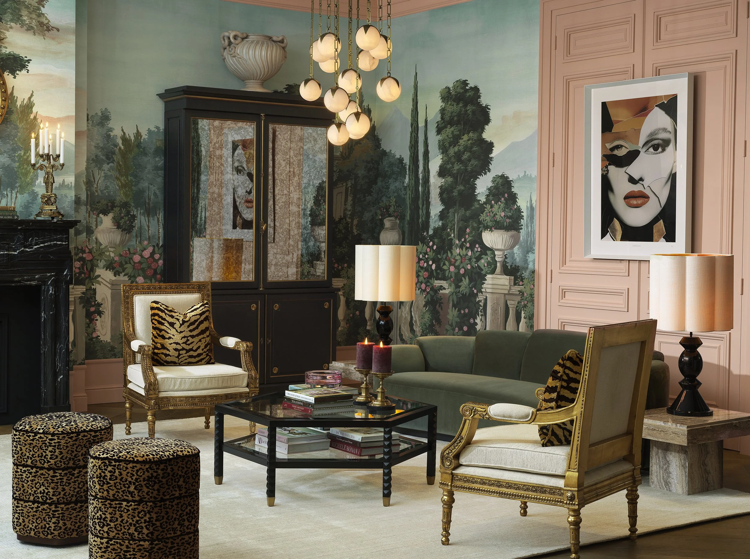 A living room with a mural of a landscape featuring mountains and trees on the wall. The room has a black wardrobe, a framed portrait of a woman with bold makeup, and classic-style chairs with gold accents. There is a chandelier hanging from the ceil