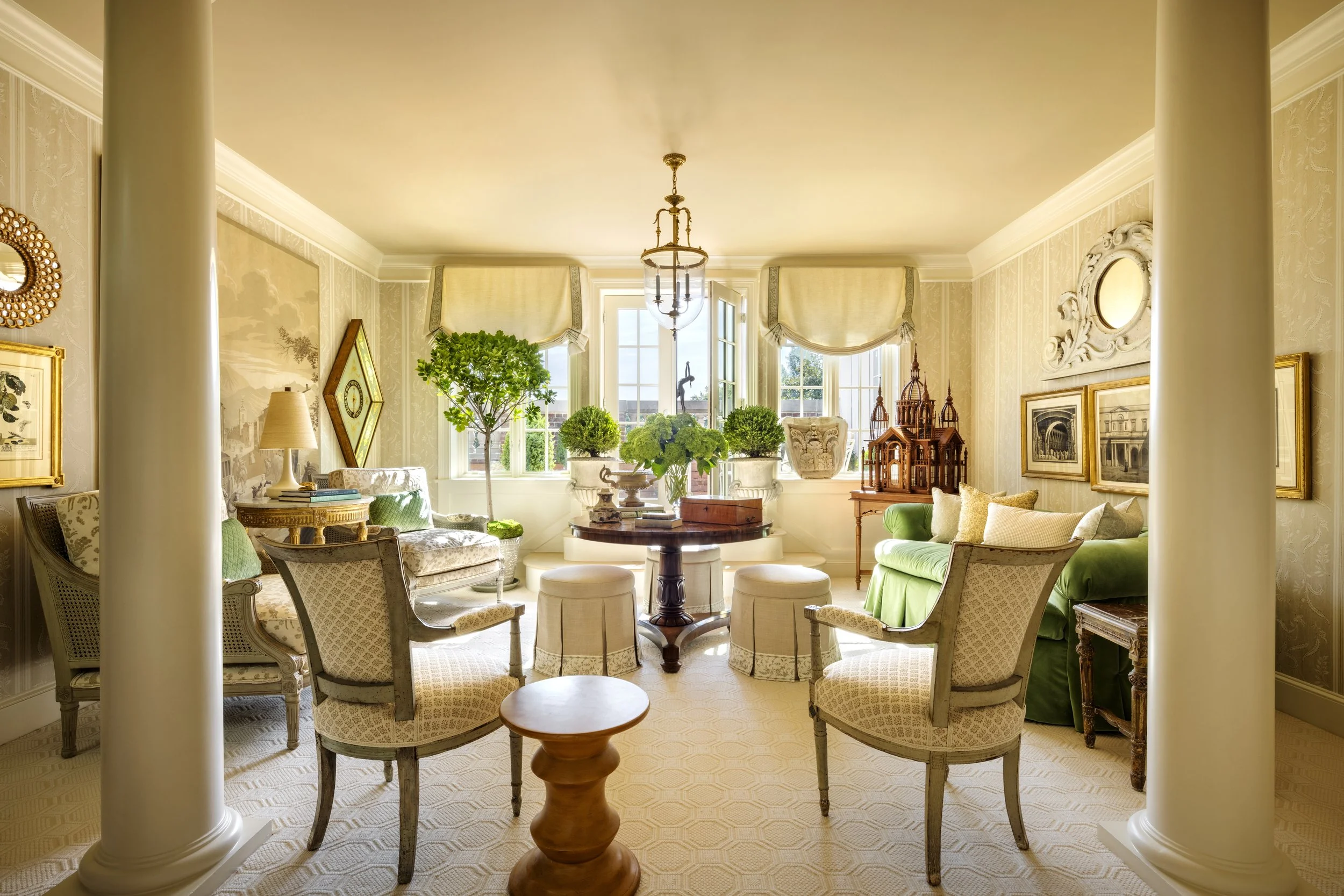 Bright and elegant living room with large windows, greenery, and classic furniture including armchairs, sofas, and a wooden coffee table.