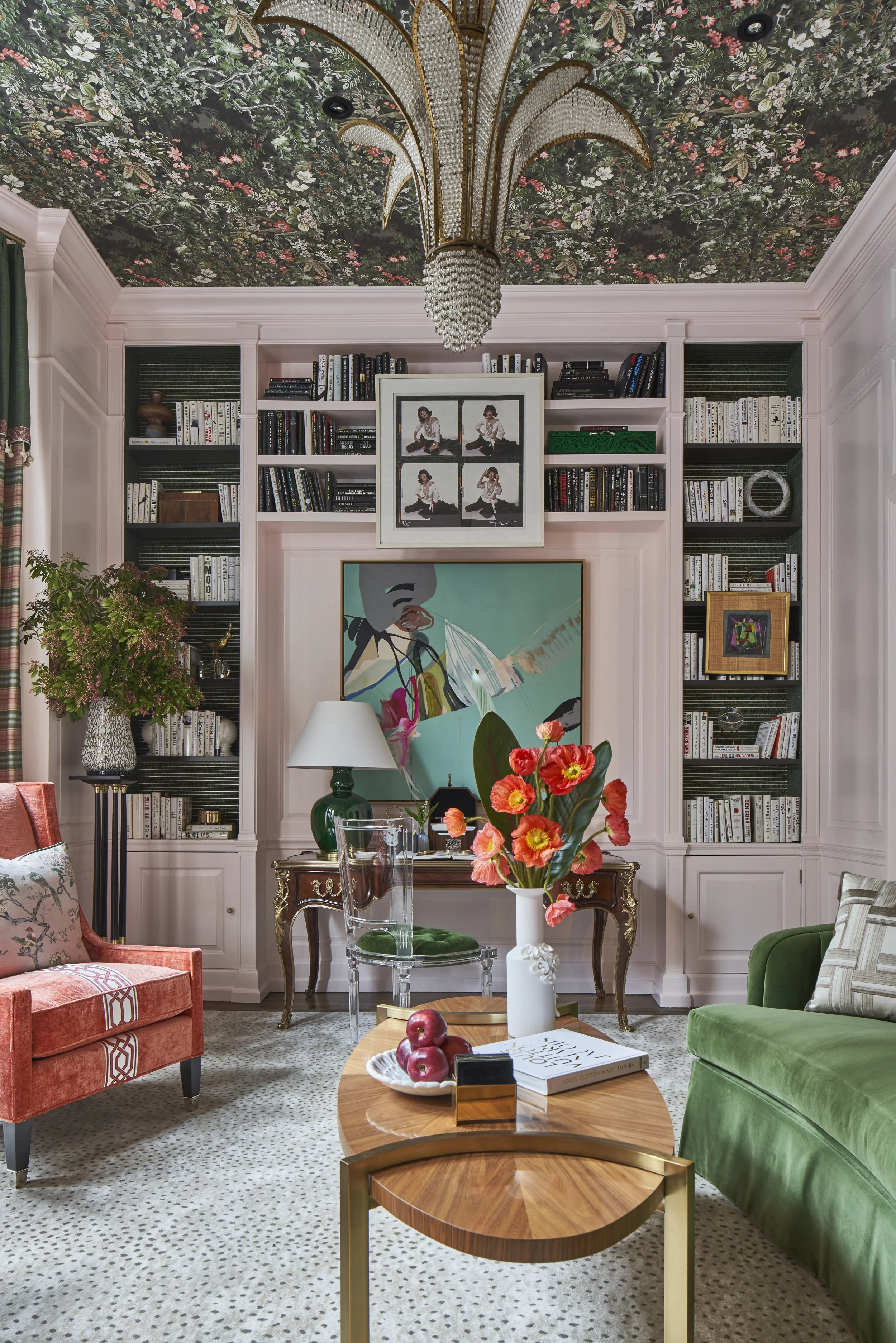 A living room with pink built-in bookshelves, a floral ceiling, a chandelier, and a variety of chairs including a green velvet chair, a pink velvet armchair, and clear acrylic chairs around a wooden table with a floral arrangement. The room also feat