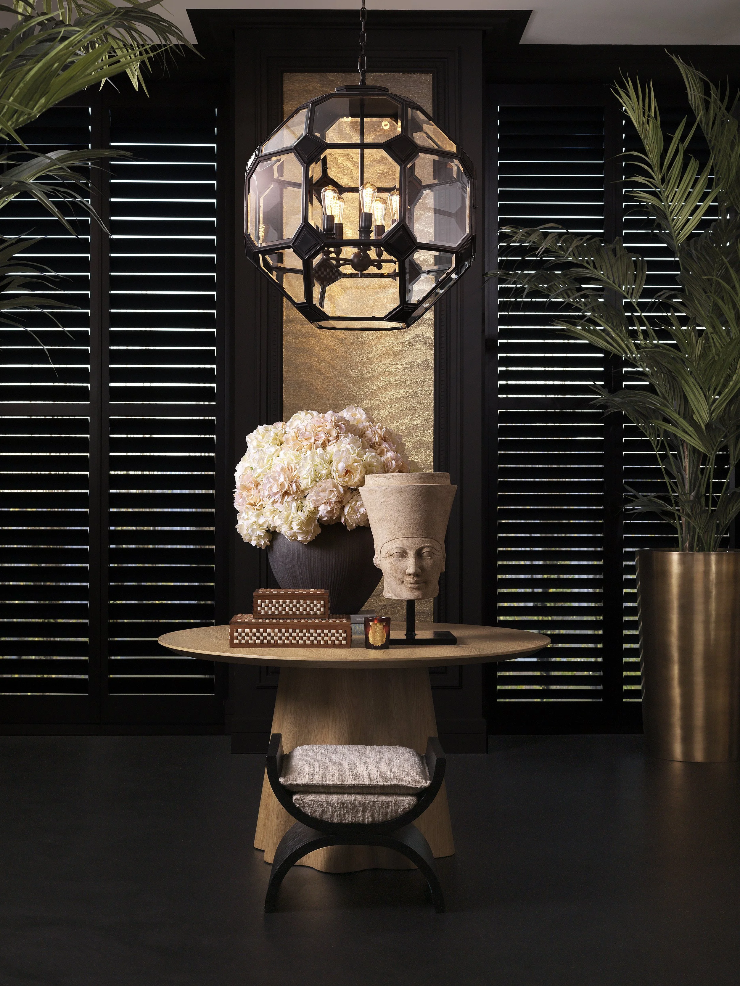Interior decor with a round wooden table, a large floral arrangement, a human head sculpture with a plant on top, a small wooden box, a lit candle, a black seating stool, a black and gold vase with a tall plant, black shutters, a gold textured wall, 