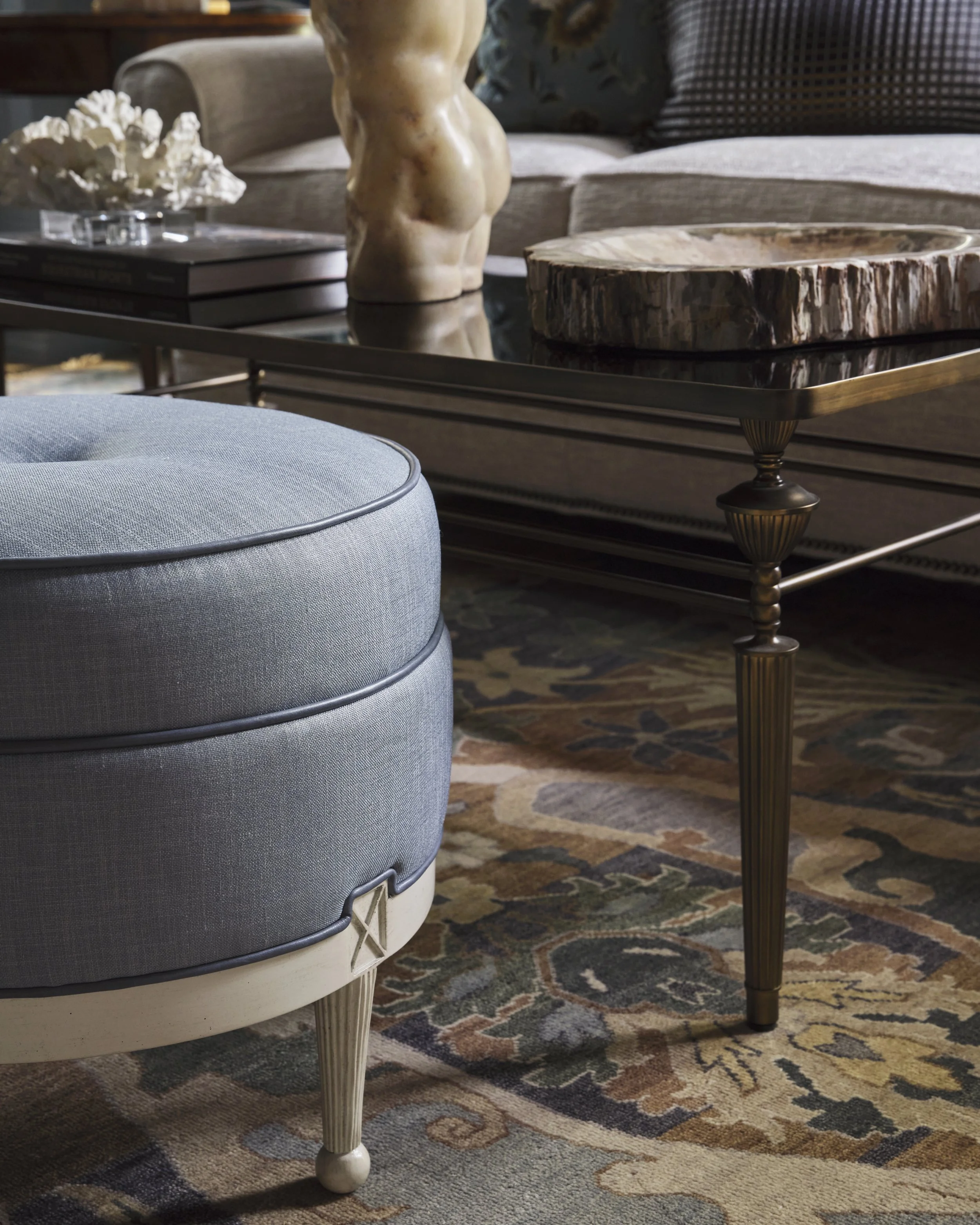 Close-up of a modern living room with a light blue upholstered ottoman, a marble-topped coffee table with a decorative sculpted bronze base, a sofa with striped and checkered pillows, a wooden sculpture of a nude female torso, and a patterned area rug.