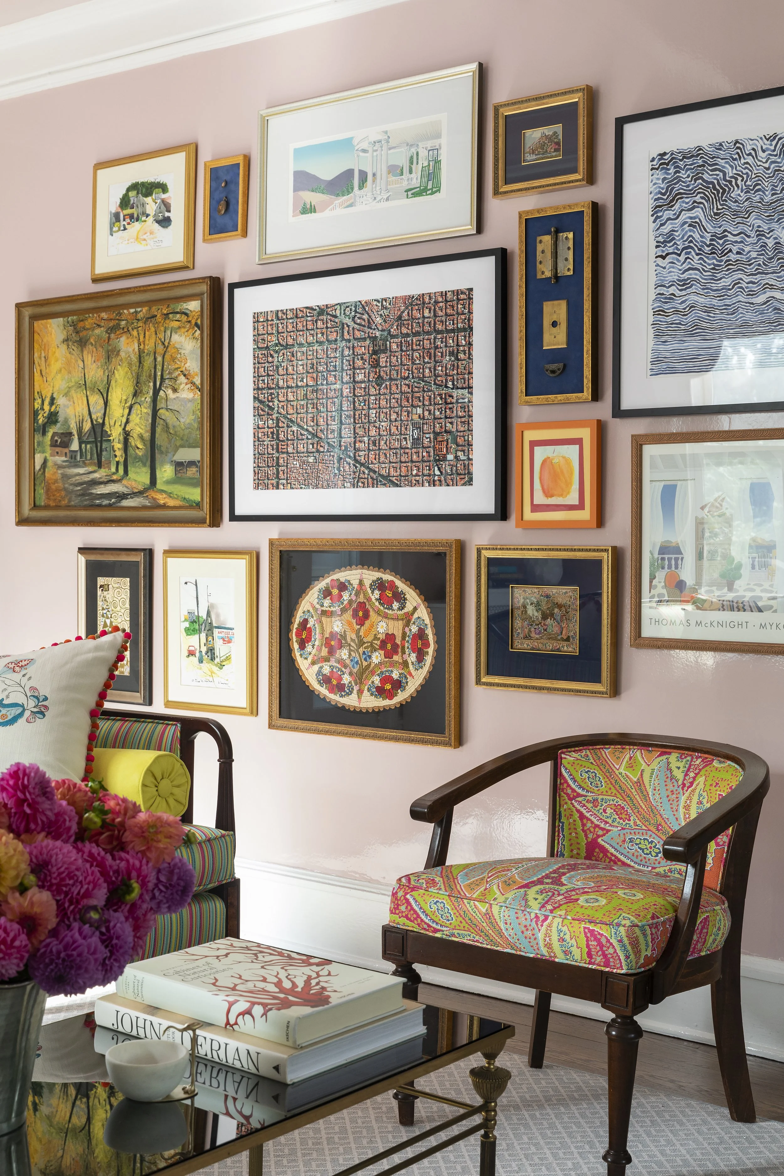 Living room with pink wall decorated with various framed artwork, colorful armchair, wooden chair, flowers, and books.