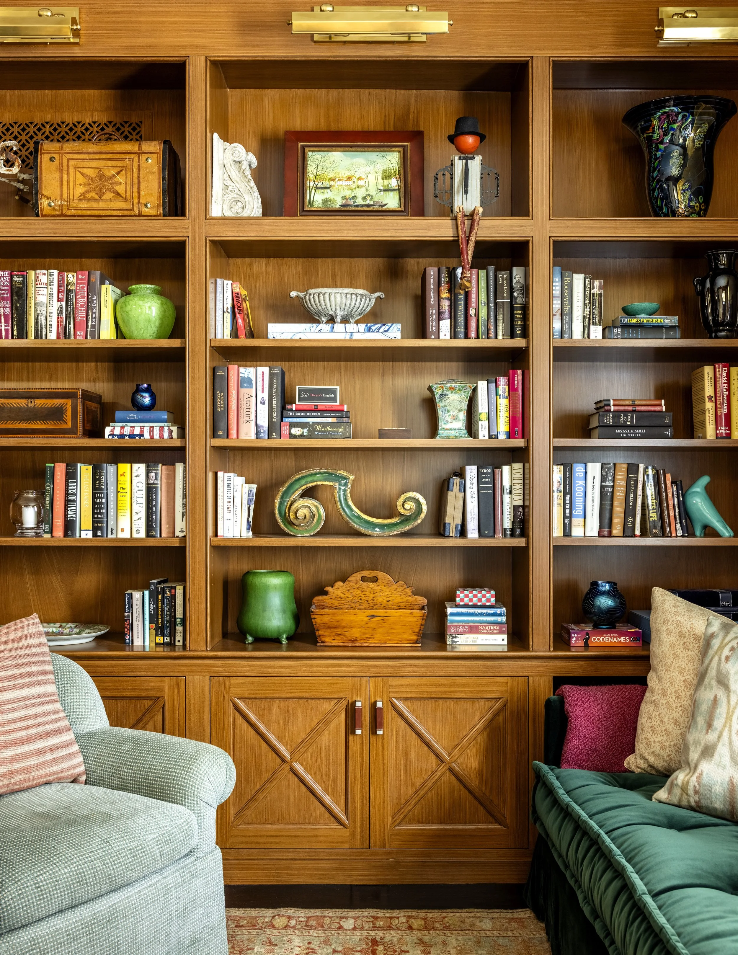 A wooden bookshelf with various decorative items and books, flanked by a light blue armchair with a pink pillow on the left and a dark green sofa with pillows on the right.