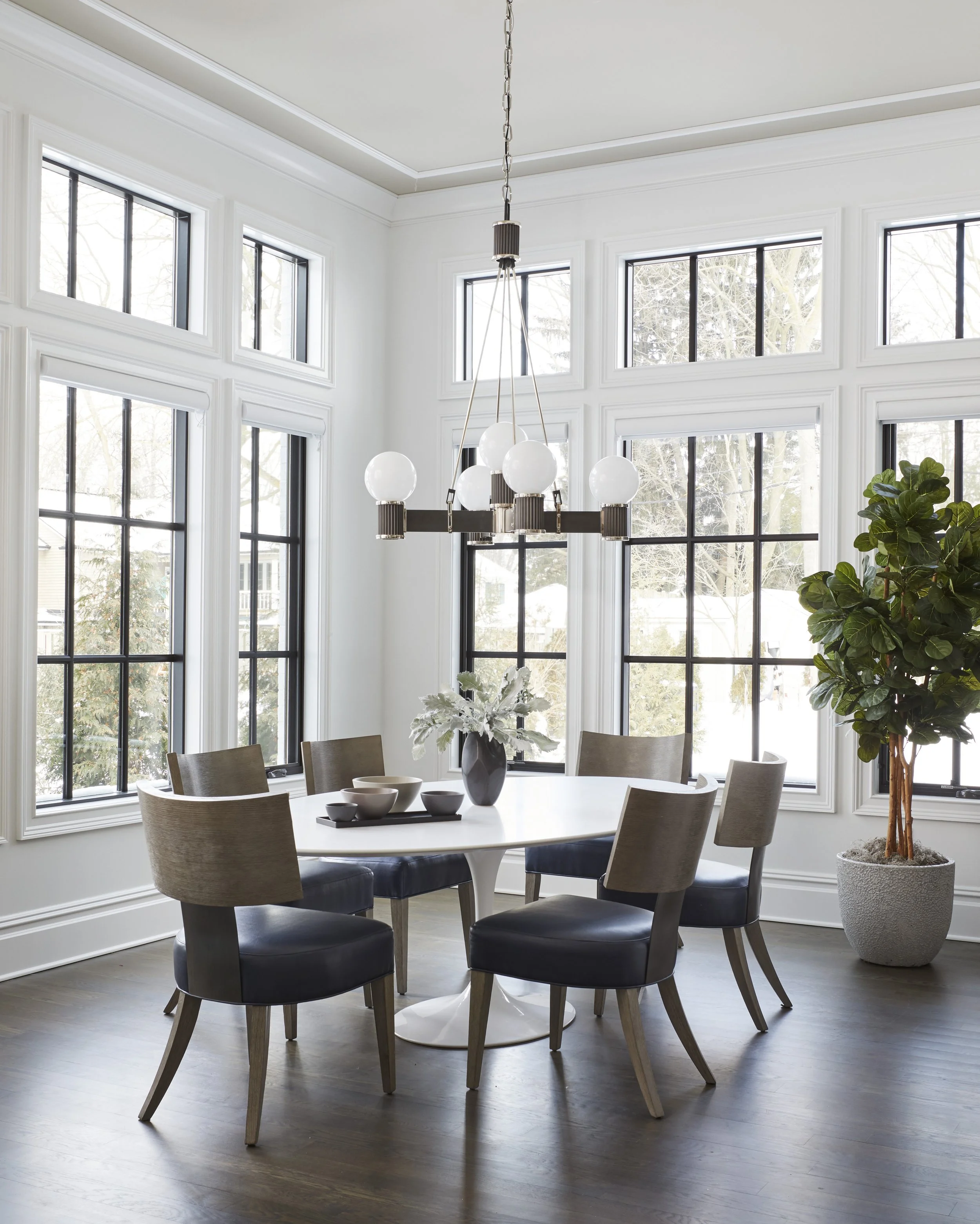 Modern dining room with large windows, a white round table, six chairs with beige and black upholstery, a black chandelier with white globe lights, a black vase with white flowers, and a large potted plant, with snow visible outside.
