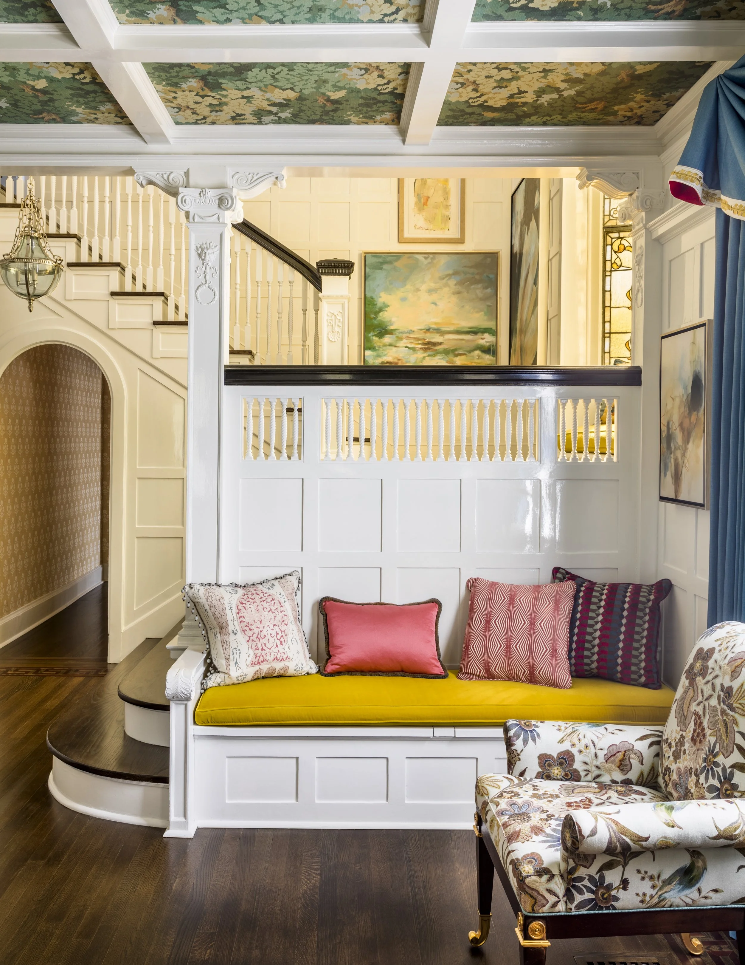 Living room with dark hardwood floors, cream-colored walls, and a staircase with detailed woodwork. A yellow bench with five decorative pillows is in front of a white wainscoting wall with paintings. A floral-patterned armchair with gold accents is p