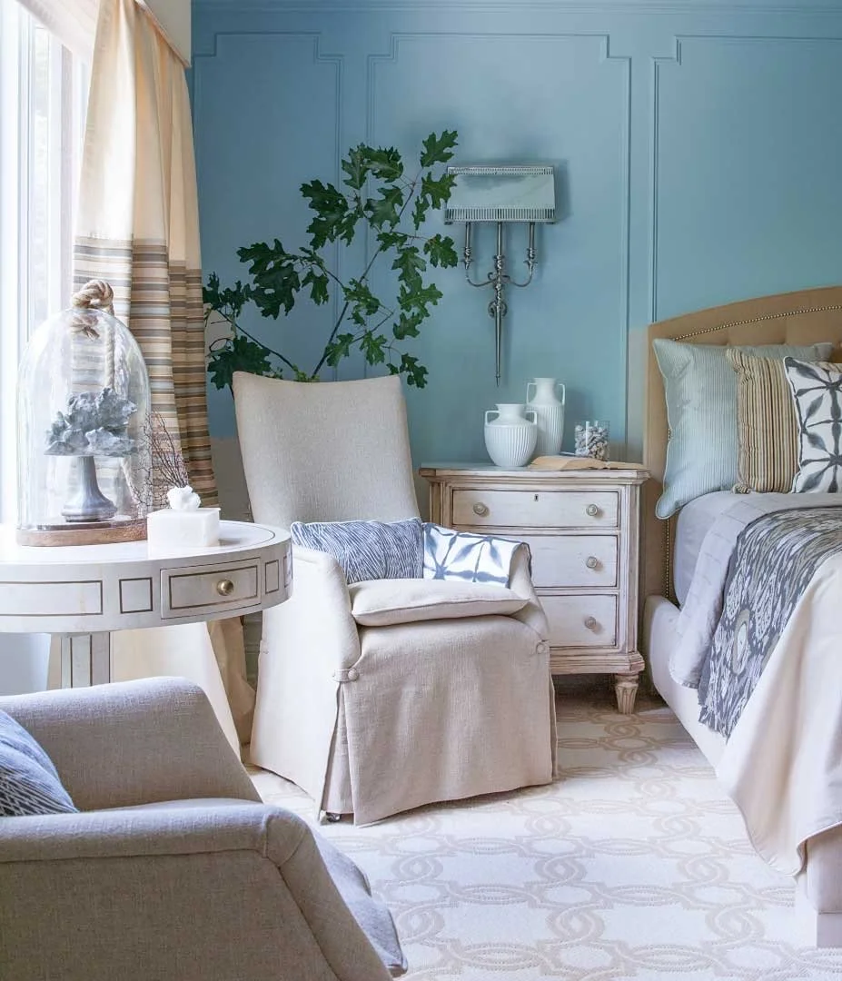 A cozy bedroom with a light blue wall, a large green plant, a nightstand with white vases and a jar, a bed with beige and patterned pillows, an armchair with a pillow and a blanket, and a window with cream curtains.