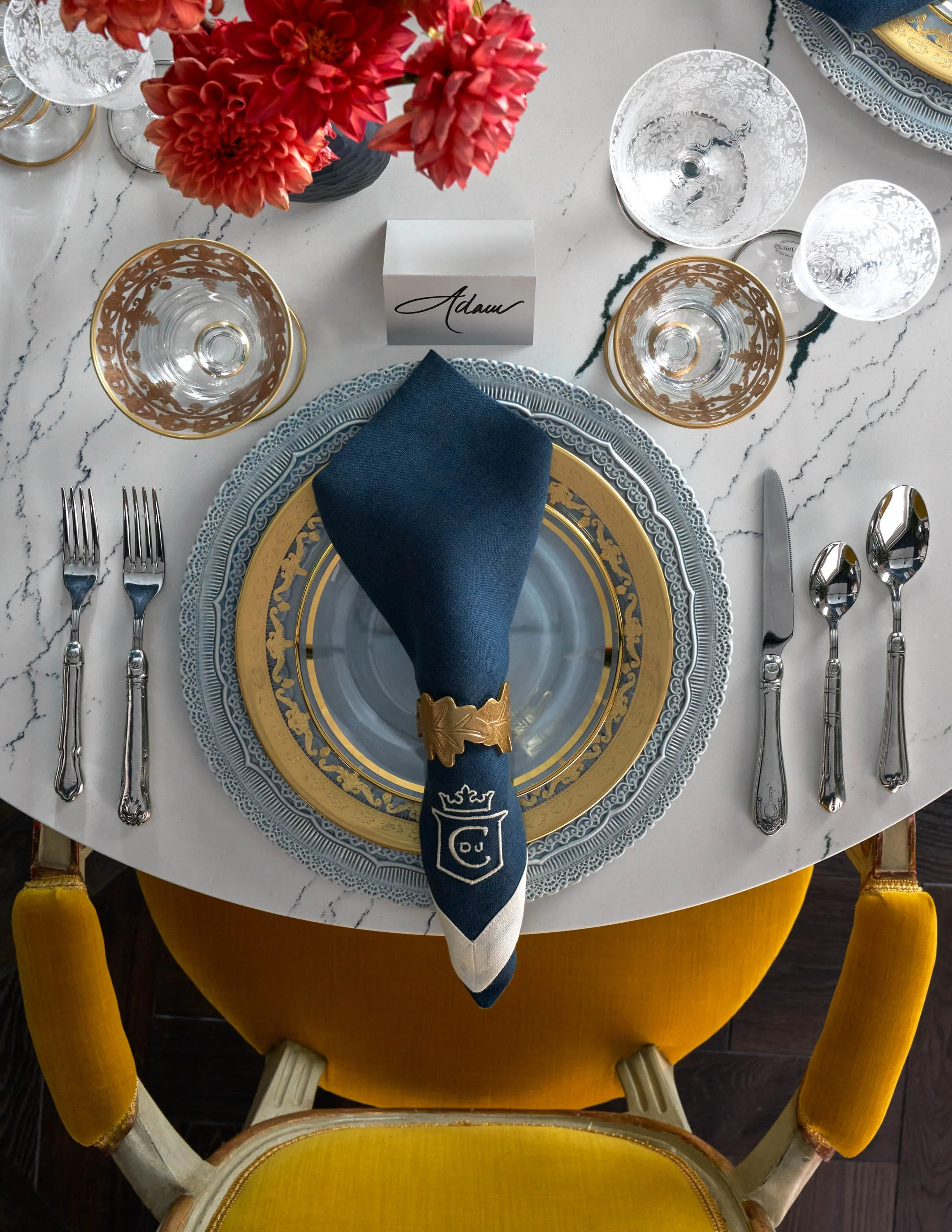 A top-down view of a table setting with elegant glassware, a gray and gold charger plate, a folded navy blue napkin with gold accents, and a place card with the name 'Adam'. There is a floral centerpiece with pink and red flowers and a yellow chair w