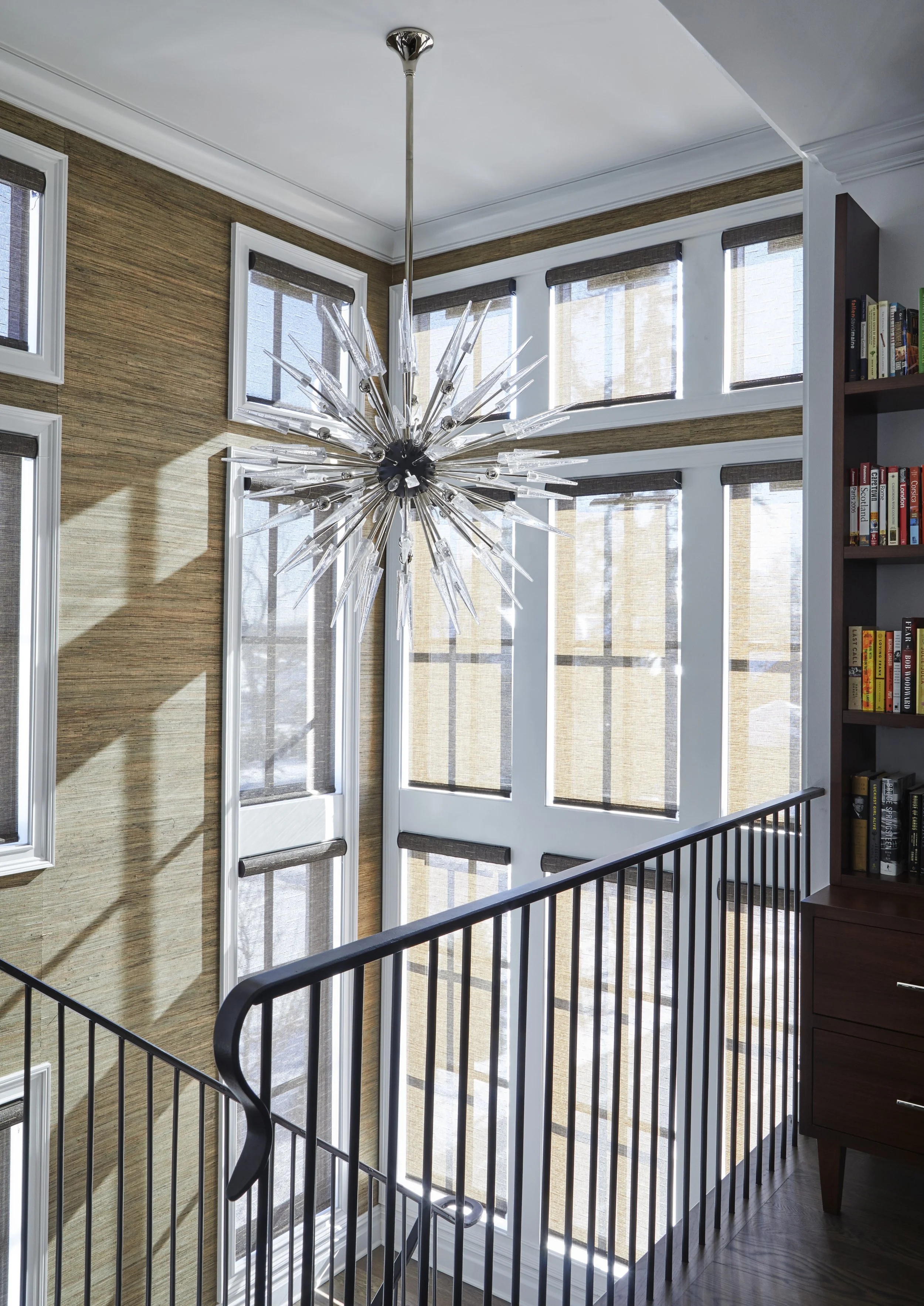Interior view of a staircase with a chandelier hanging from the ceiling, multiple large windows with shades, and a bookshelf on the right side.