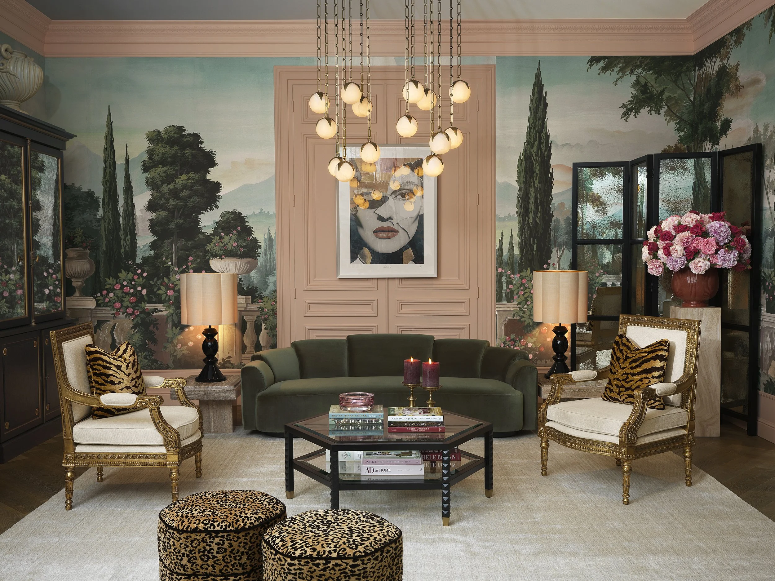 Living room with a green velvet sofa, white armchairs with gold trim, a black coffee table with books and candles, a large floral arrangement, a chandelier with multiple globes, and a mural of scenic landscape with trees and mountains on the walls.