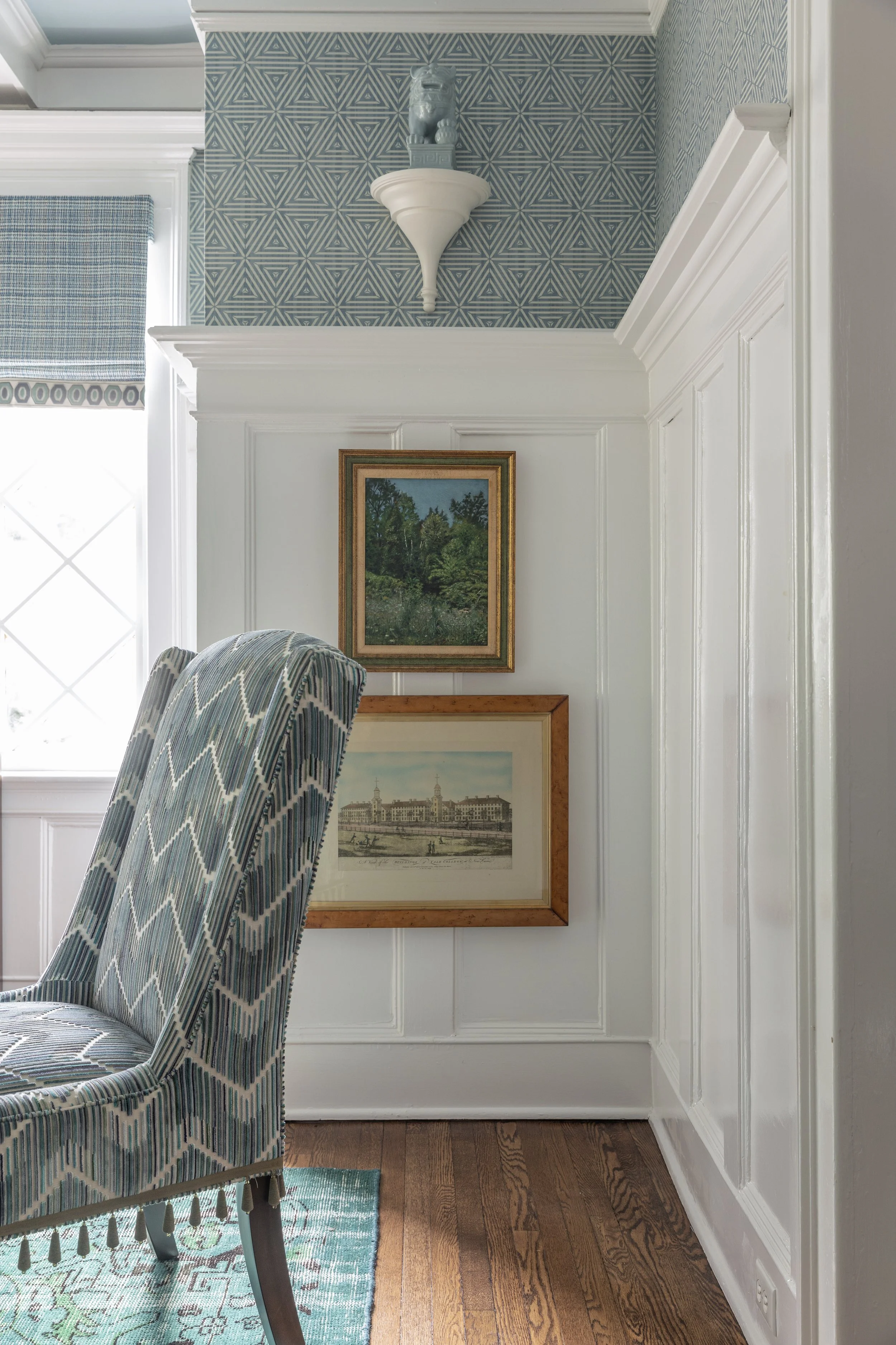 A corner of a dining room with a patterned armchair in the foreground, featuring a zigzag design in shades of blue, green, and beige. The wall behind has white vertical paneling with two framed pictures: one of a landscape with green trees and a buil